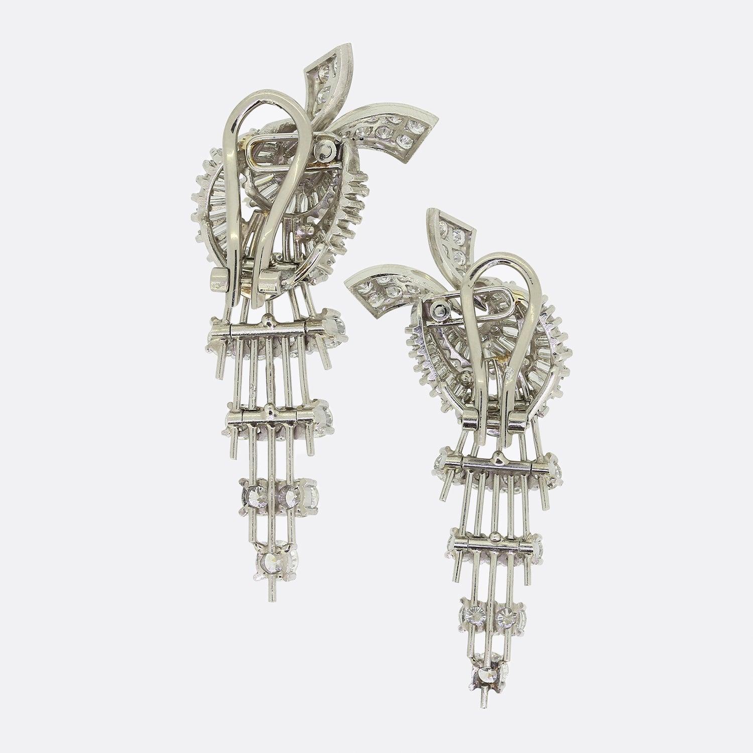 Here we have an exceptionally radiant pair of retro diamond earrings from the 1950s. Both open frames showcase graduating groups of round brilliant cut diamonds which hang freely below a ribbon motif pervaded with baguette cut diamonds and matching