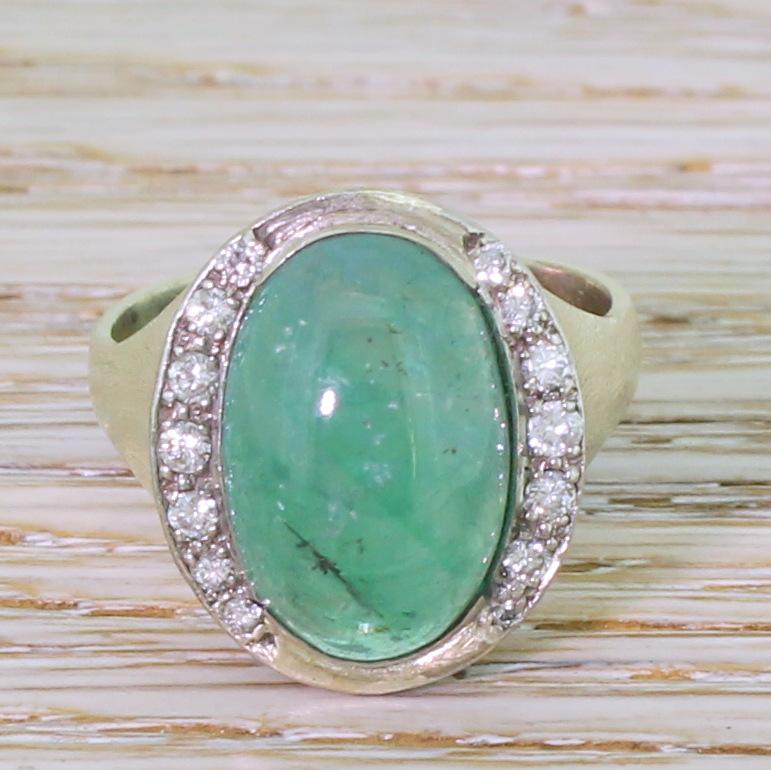 A bold and dramatic vintage emerald ring. The oval shaped cabochon emerald is bright, light blueish green – the colour of which is emphasised by the two crescents of seven (fourteen total) round brilliant cut diamonds that border the long sides of