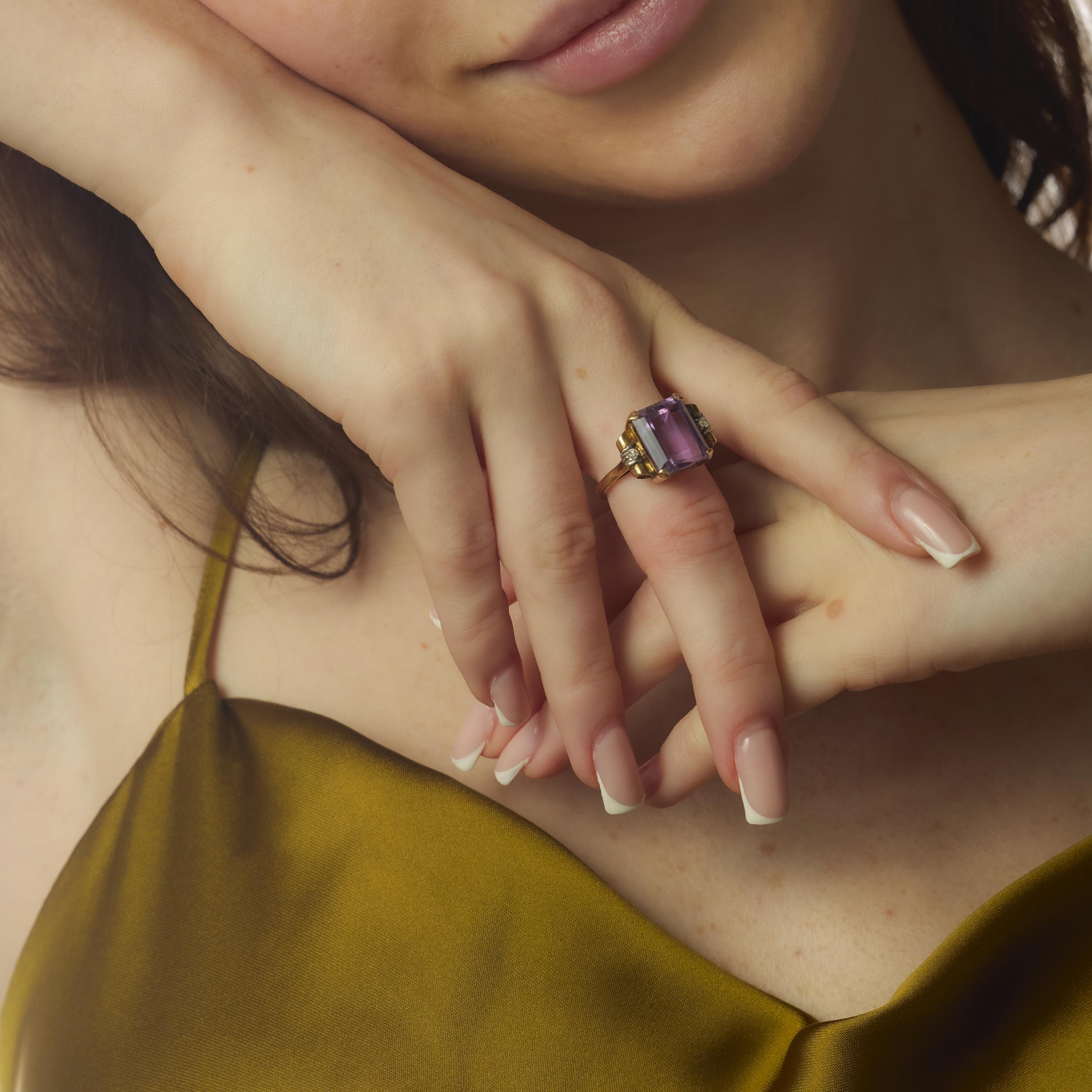 Cette bague rétro des années 1940 est ornée d'une vibrante améthyste de 6,90 carats de taille émeraude, dont la surface facettée rayonne d'une riche teinte violette. La pierre centrale est flanquée de deux petits diamants taille unique, qui ajoutent