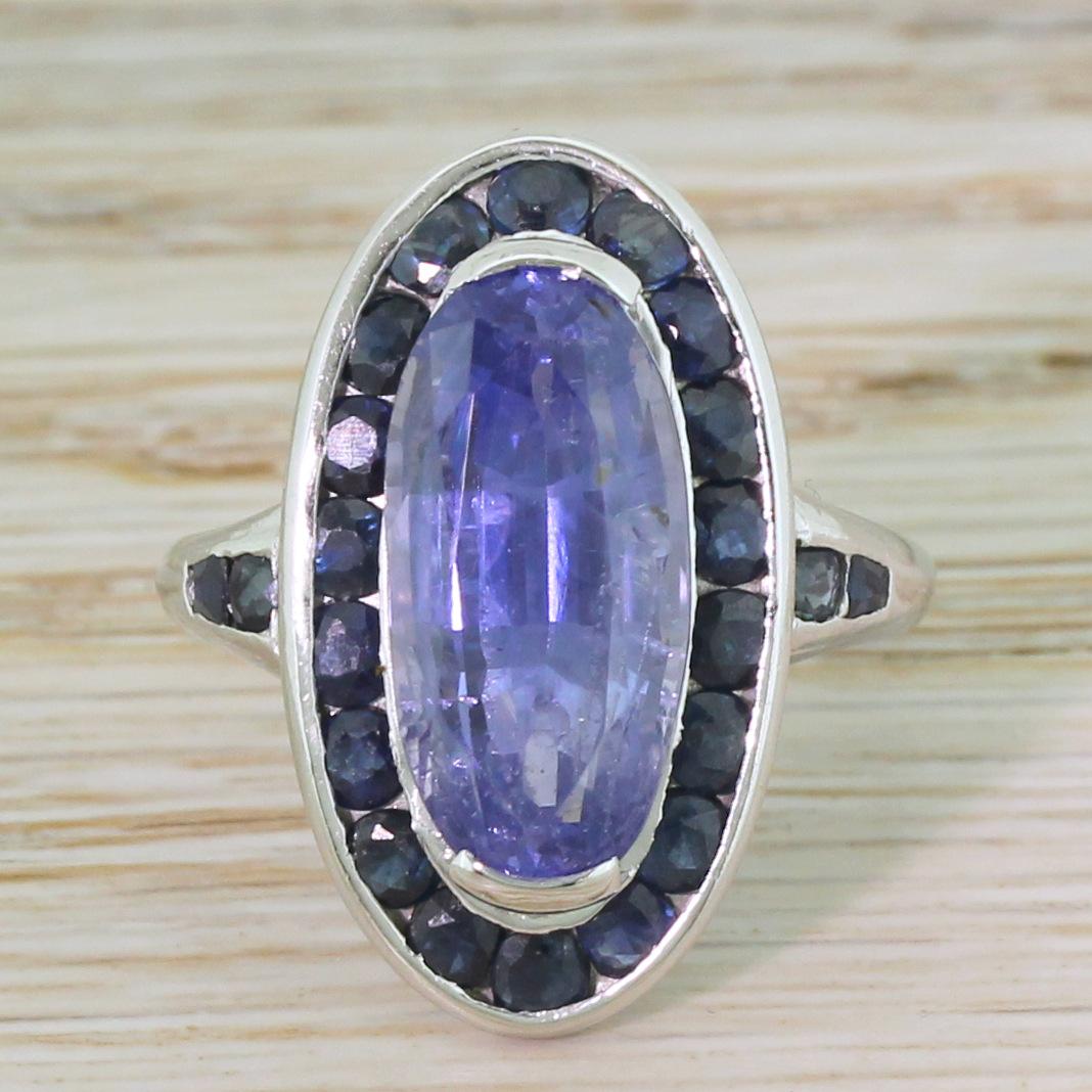 A wonderful retro ring, dressing in blue. The oval cut centre sapphire – certified as natural, unheated and originating from Ceylon – displays a glowing cornflower hue. Eighteen rich, royal blue sapphire are channel set in the surround, with ornate