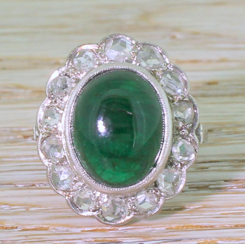 A hypnotically beautiful cabochon emerald ring. The large, high domed centre stone is a verdant, glowing, bottle green and is rubover and milgrain set in white gold with fourteen white and bright rose cut diamonds in the surround. Striking detailing