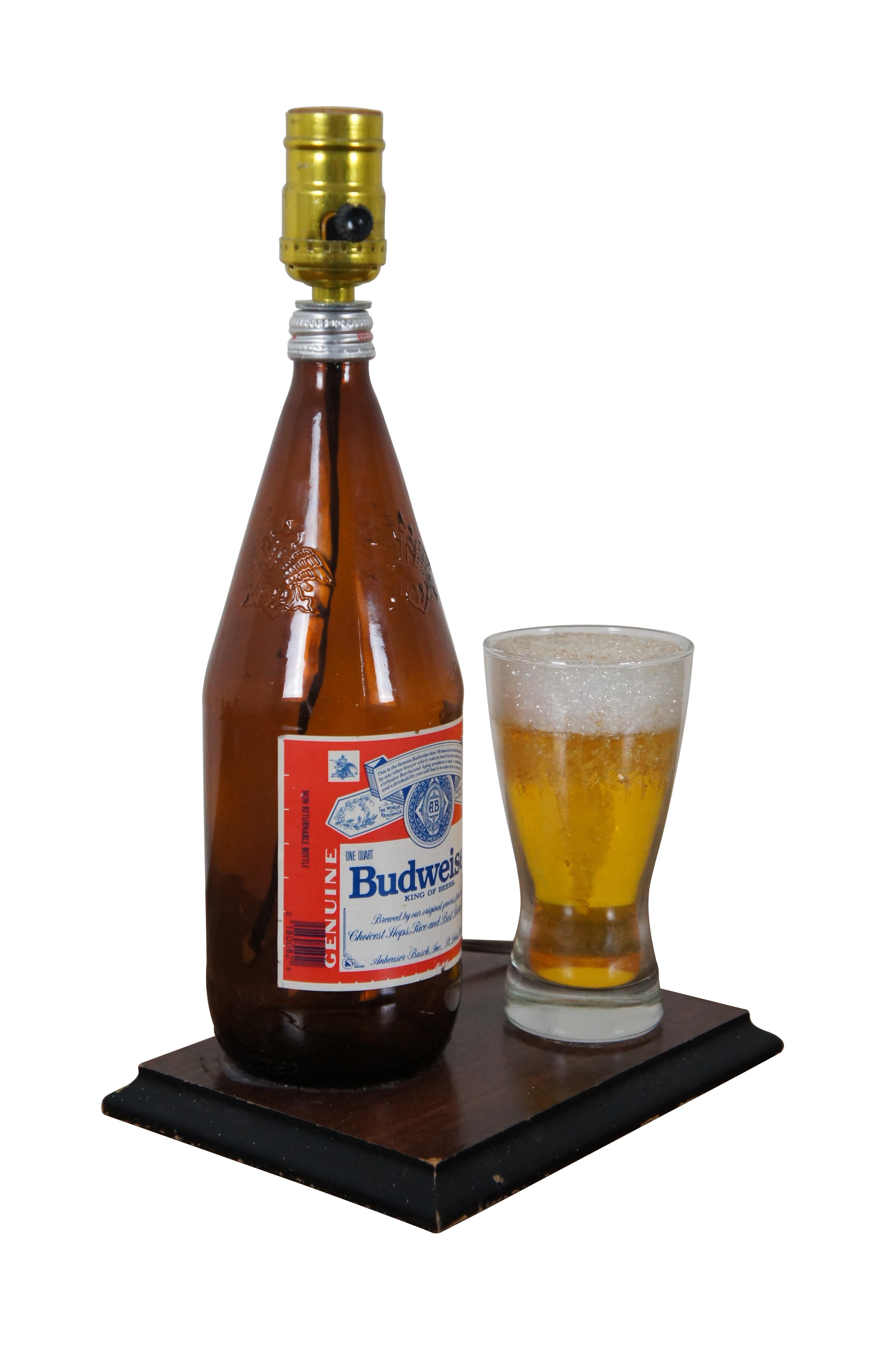 Vintage beer bottle and drinking glass table lamp, crafted from a repurposed Budweiser beer bottle, mounted on a wooden plaque for a base, next to a beer glass filled with amber colored resin “beer” and topped with a frothy Styrofoam “head.”