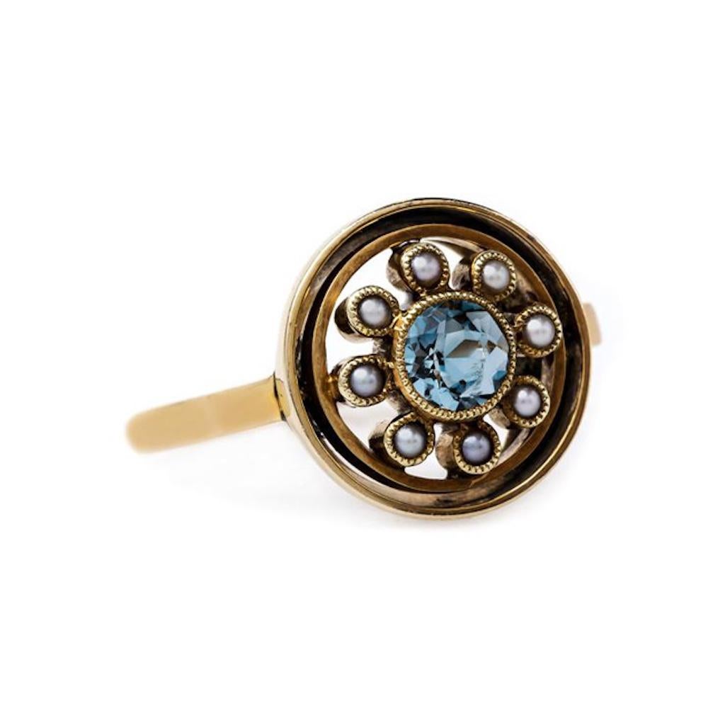 This is a unique and whimsical Retro era (circa 1940) 14k yellow gold ring. This fun ring centers a bezel set Round cut aquamarine gauged at 5mm totaling approximately 0.40ct. The ocean blue center stone is bordered by a halo of gray seed pearls,