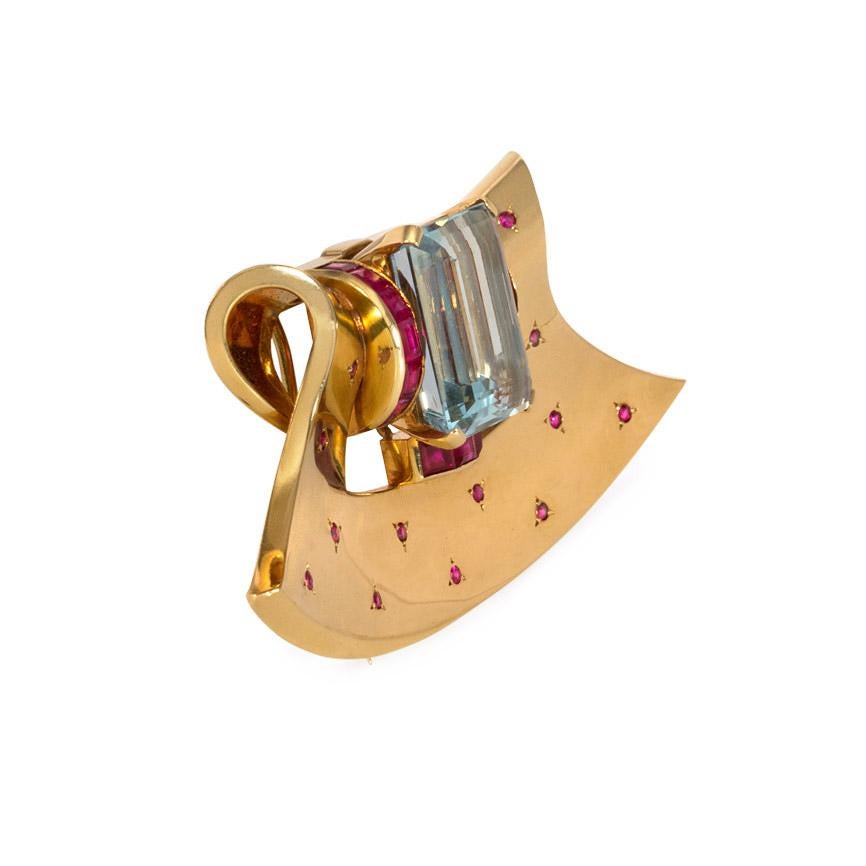 Eine Retro-Gold-Clip-Brosche mit fächerförmigem Design, besetzt mit einem großen zentralen Aquamarin, der an drei Seiten von Rubinen im Kaliberschliff umrahmt ist, akzentuiert mit Rubinen im Rundschliff, in 18k.  Der Aquamarin misst 19,00 x 14,75 x