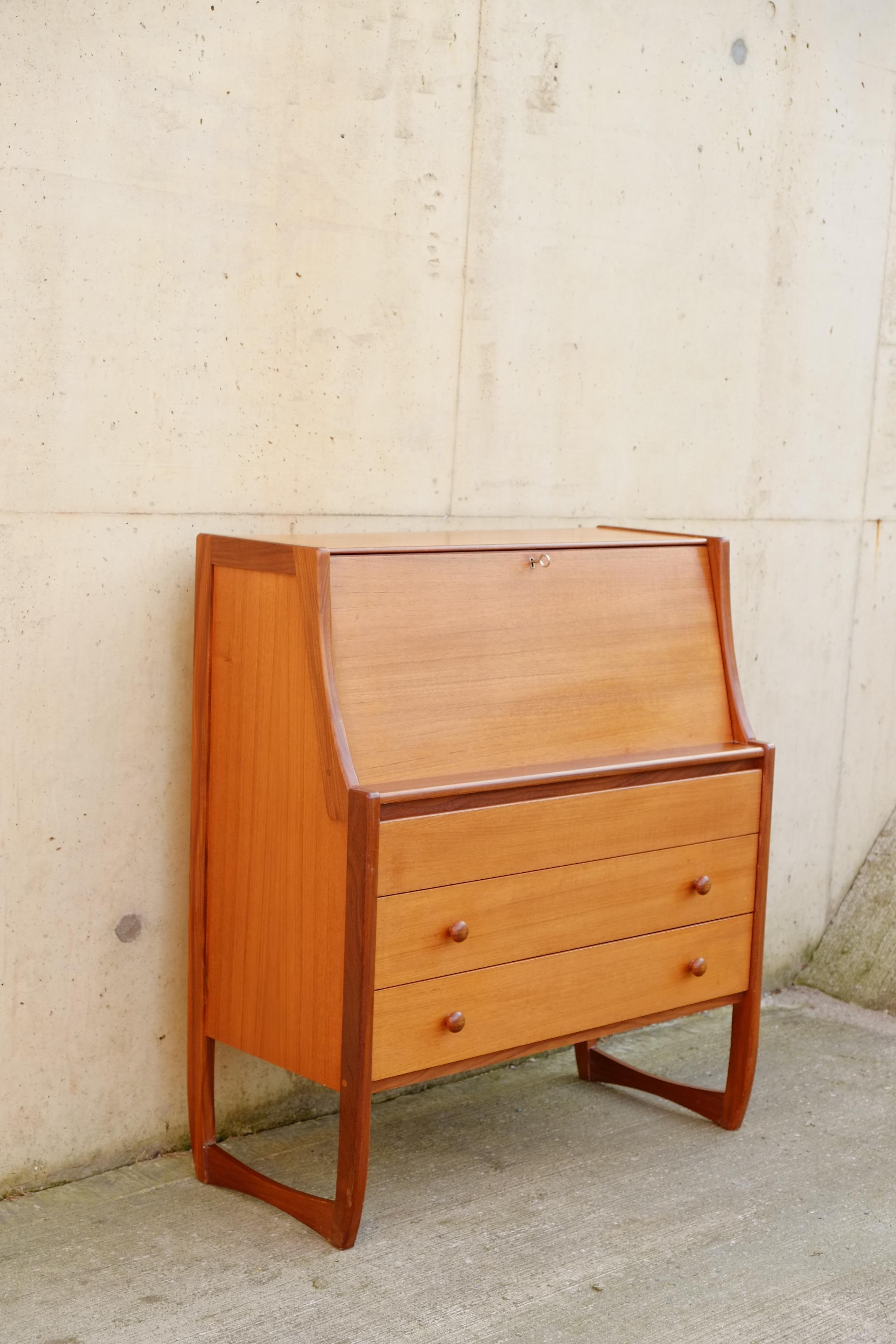 A cute retro teak bureau desk with three drawers. Lovely sculpted legs in solid teak, three clean drawers with white carcasses and a simple drop down desk with key. The desk has multiple compartments inside for letters and paperwork. Cute small