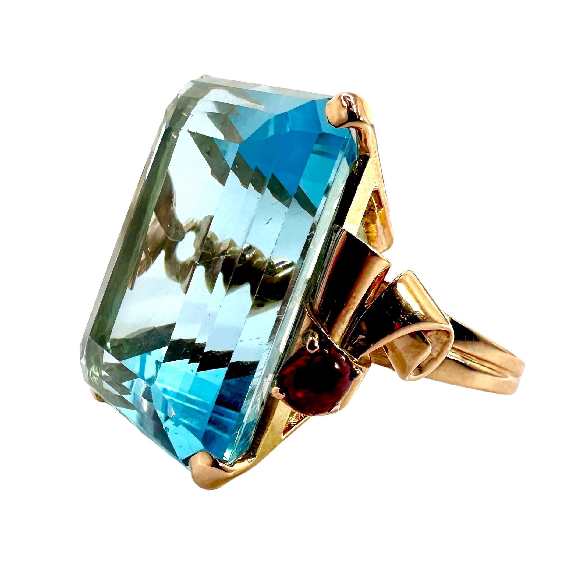 A stunning Retro ring, in 14K pink gold, centered around one emerald cut aquamarine weighing approximately 51.35 carats. This beautifully detailed ring has rounded scroll motifs on the shoulders set with cabochon rubies for an amazing contrast of