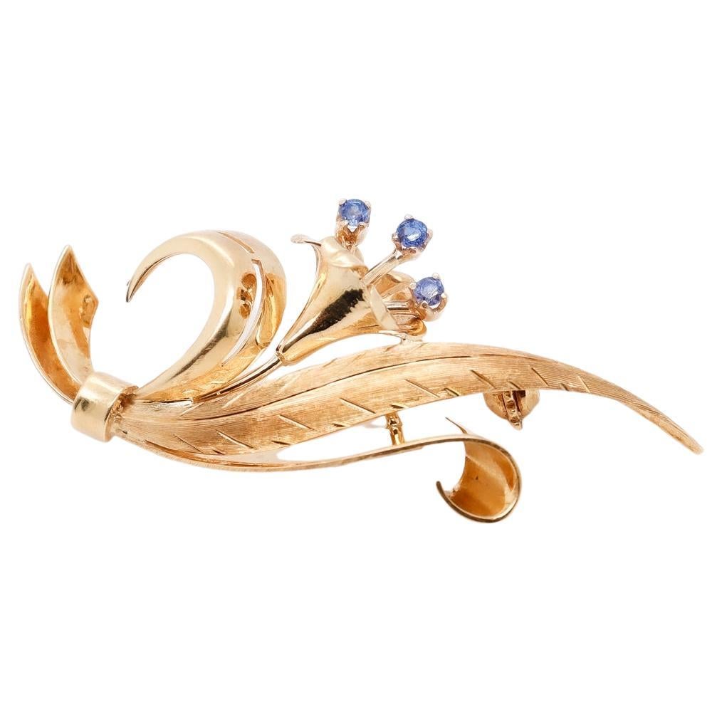 A fine retro flower brooch.

In 18k gold.

By Tiffany & Co.

With a stylized flower (likely a Lily) set with small blue topaz gemstones to its pistils and flanked by a long textured leaf and scroll devices.

Simply a wonderful retro Tiffany & Co.