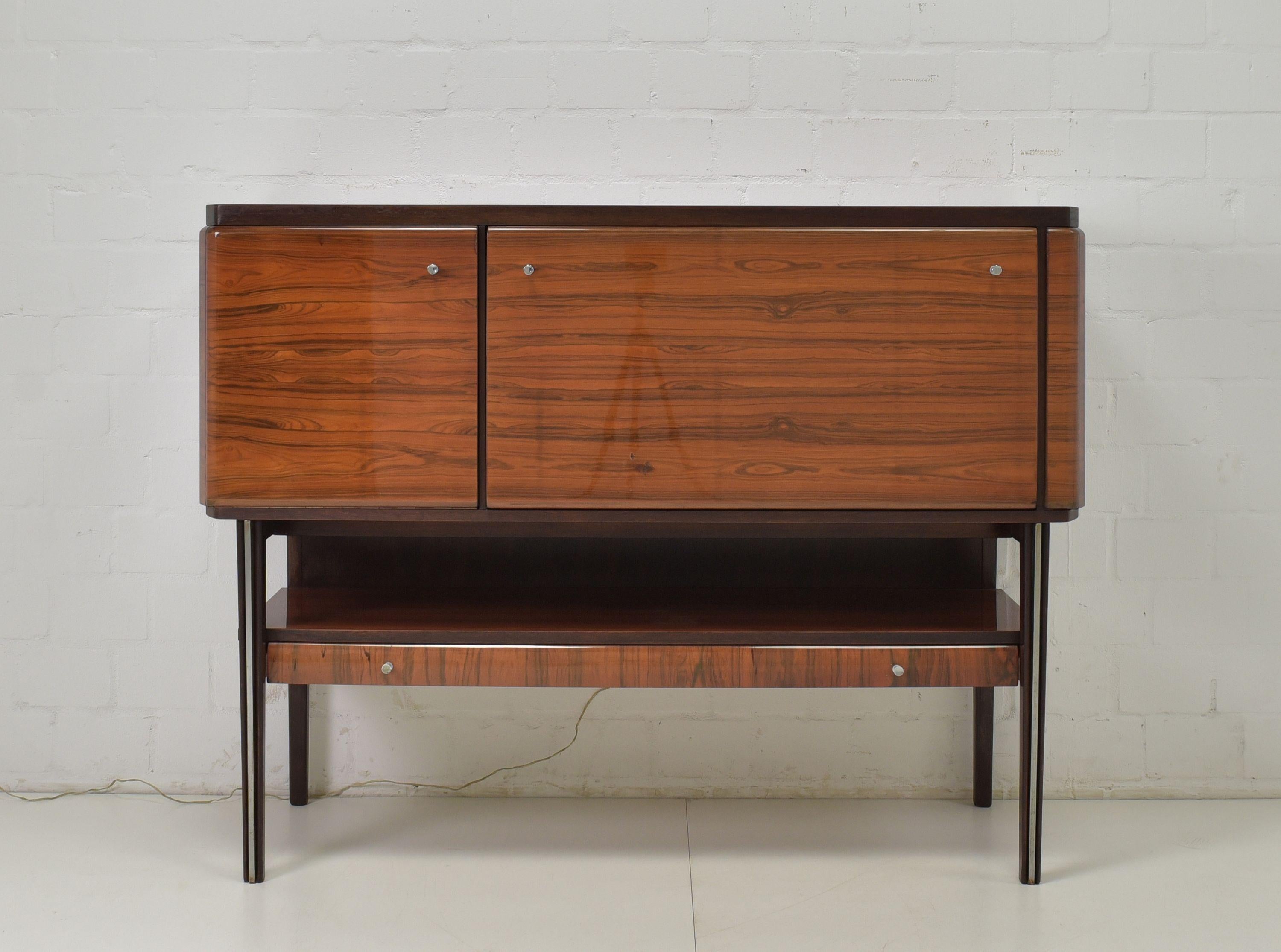 Bar cabinet highboard sideboard rosewood retro vintage around 1970 illuminated

Features:
Two-piece model
Upper part with two flaps, right side illuminated
High-legged base with an open compartment and a flat drawer
Chromed handles and chrome