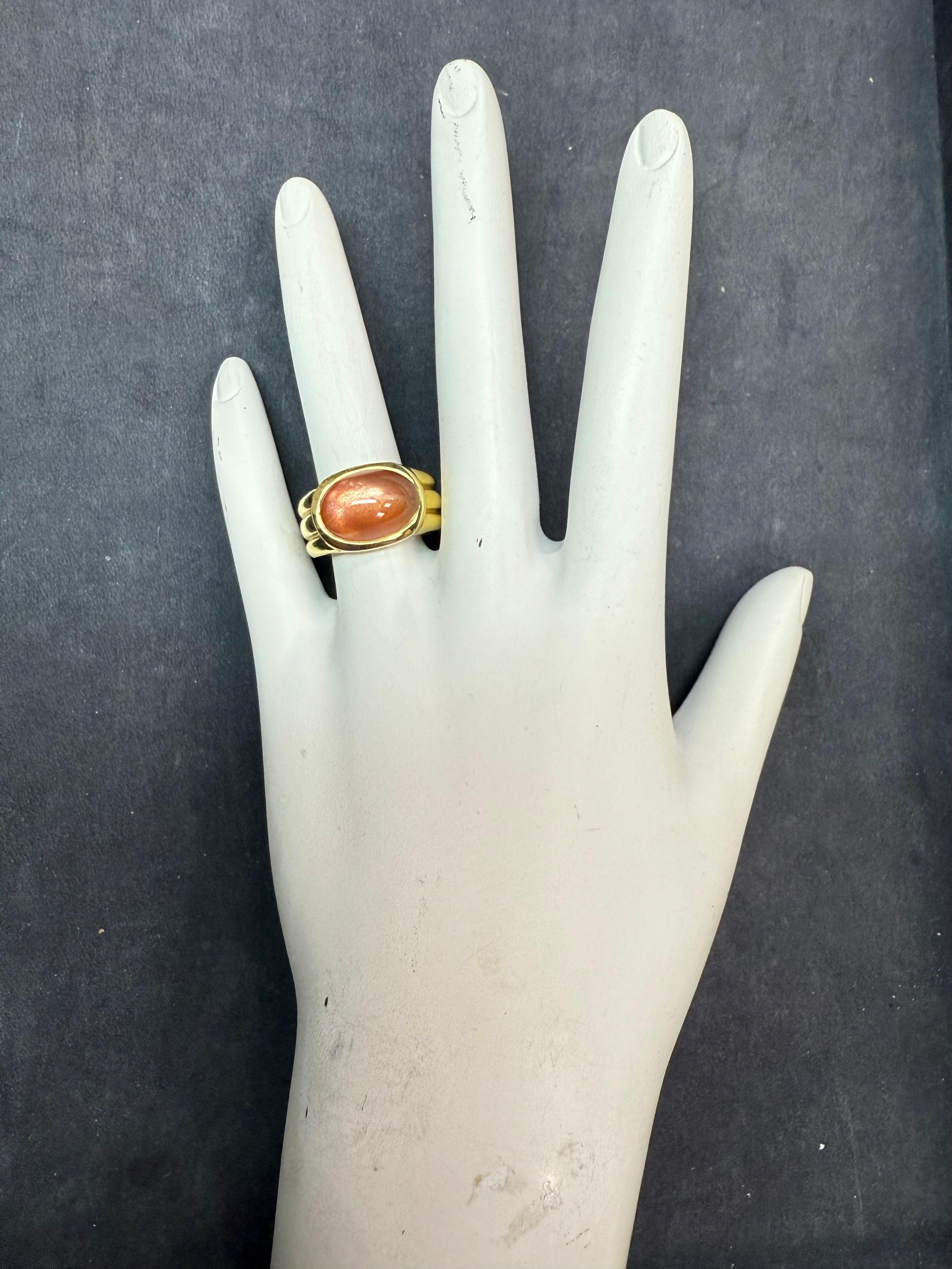 A striking oval cabochon sunstone feldspar ring set in 18K yellow gold. The center stone measures approximately 13.5 x 7 mm, showcasing the gem’s natural inner glow. The band tapers from 9.4 mm to 7.2 mm for a bold yet balanced look. Stamped 18K,