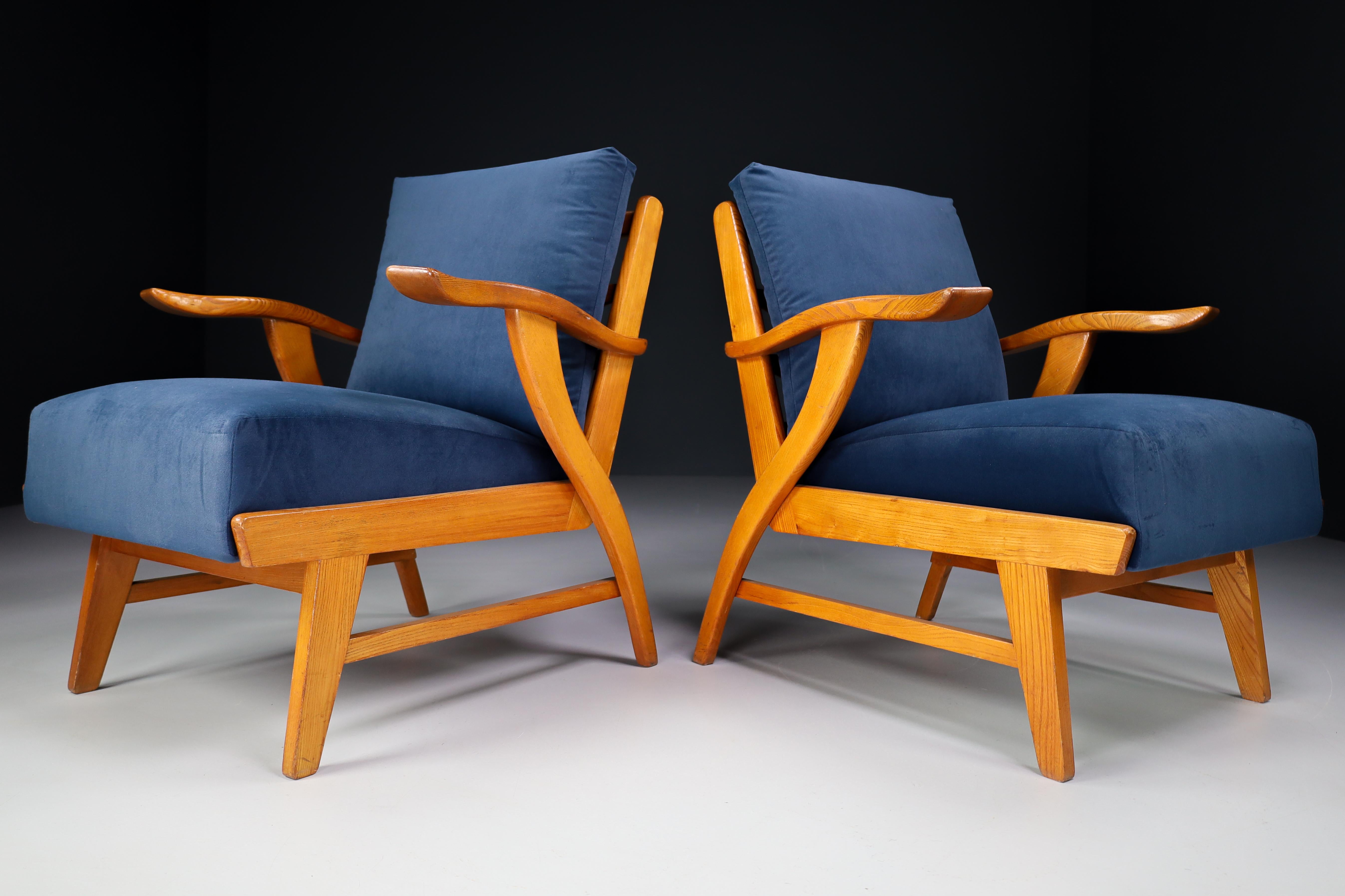 Set of two lounge chairs, in patinated solid ash and Reupholstered bleu velvet, France, 1960s. The grain of the wood is nicely visible, especially on the wide sculpted armrests. These armchairs would make an eye-catching addition to any interior