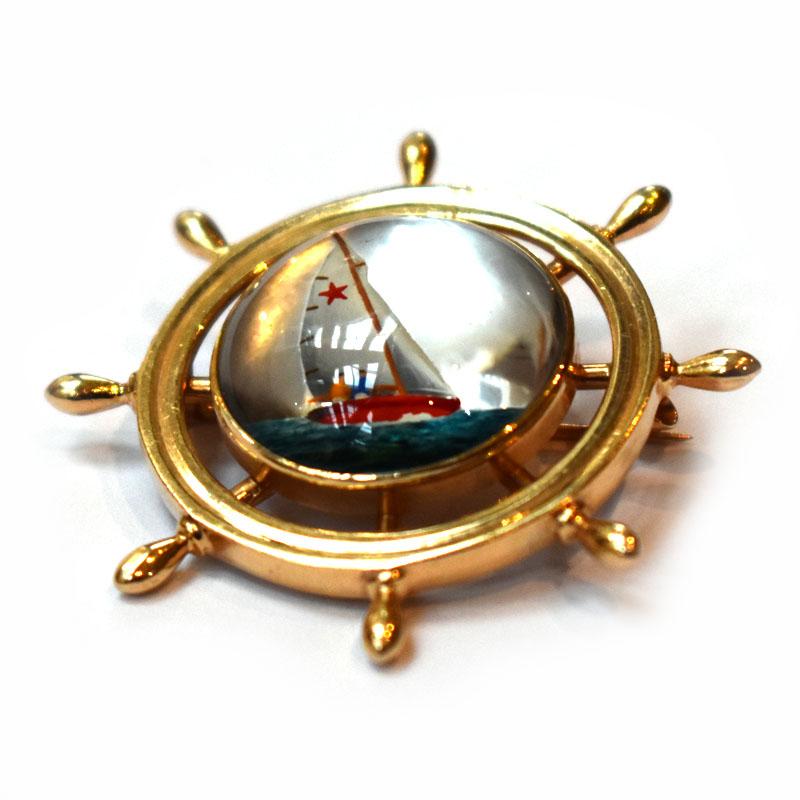 A nautical brooch featuring a reverse crystal painted with a yacht on a gold ship's wheel mount. Mounted in 14ct yellow gold, stamped 14K, circa 1970. With a stylised bow and arrow maker's mark, American.
