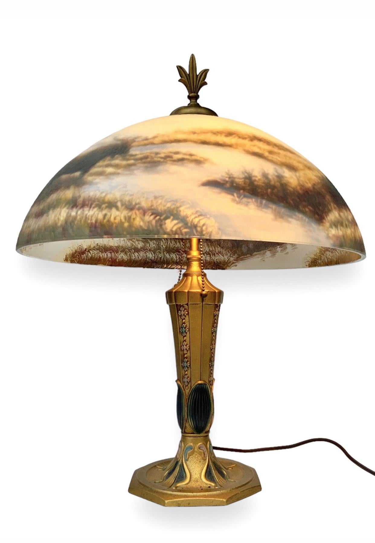 Reverse hand painted glass shade depicting Elk in their natural habitat. A small stream with tall grass and forest with mountains in the background. The metal lamp body is polychromed with gold and jewel tones. In the manner of Moe Bridges with no
