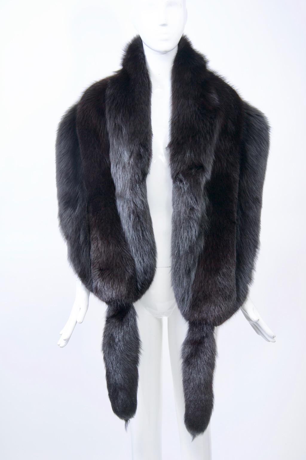 Sumptuous, full skins define this glamorous black fox shawl. Crafted of three skins that are wide and long, the shawl has rounded ends ornamented by a single tail each. Lined in black velvet with hand-hold pockets near the ends. Labelled Revillon,