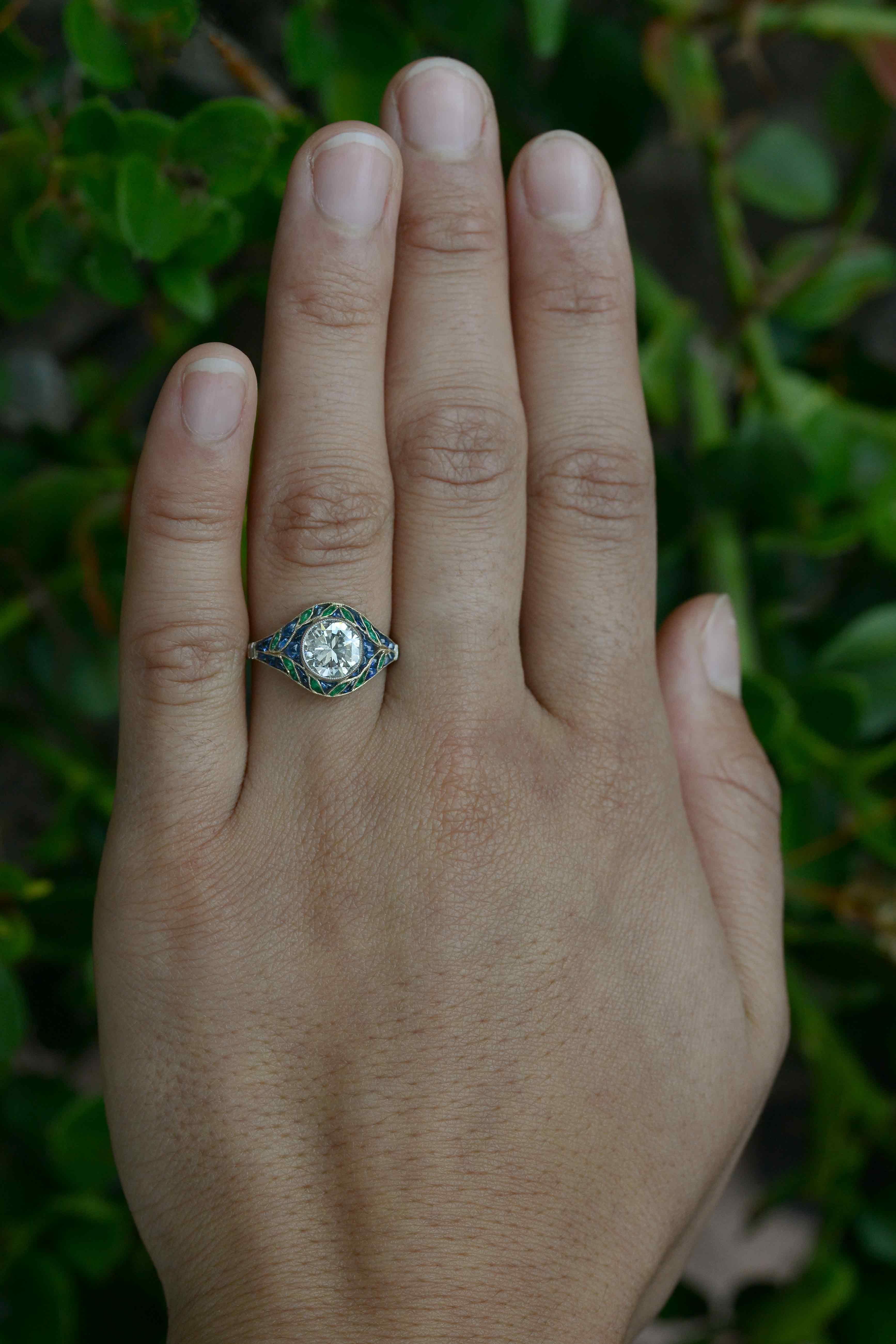 With a mesmerizing mosaic of emerald and sapphire leaves embracing a shimmering 1.41 carat round diamond, this Art Deco Style diamond engagement ring is ultra-intriguing. The floral wreath of blue and green gemstones, set in 18k yellow gold, provide