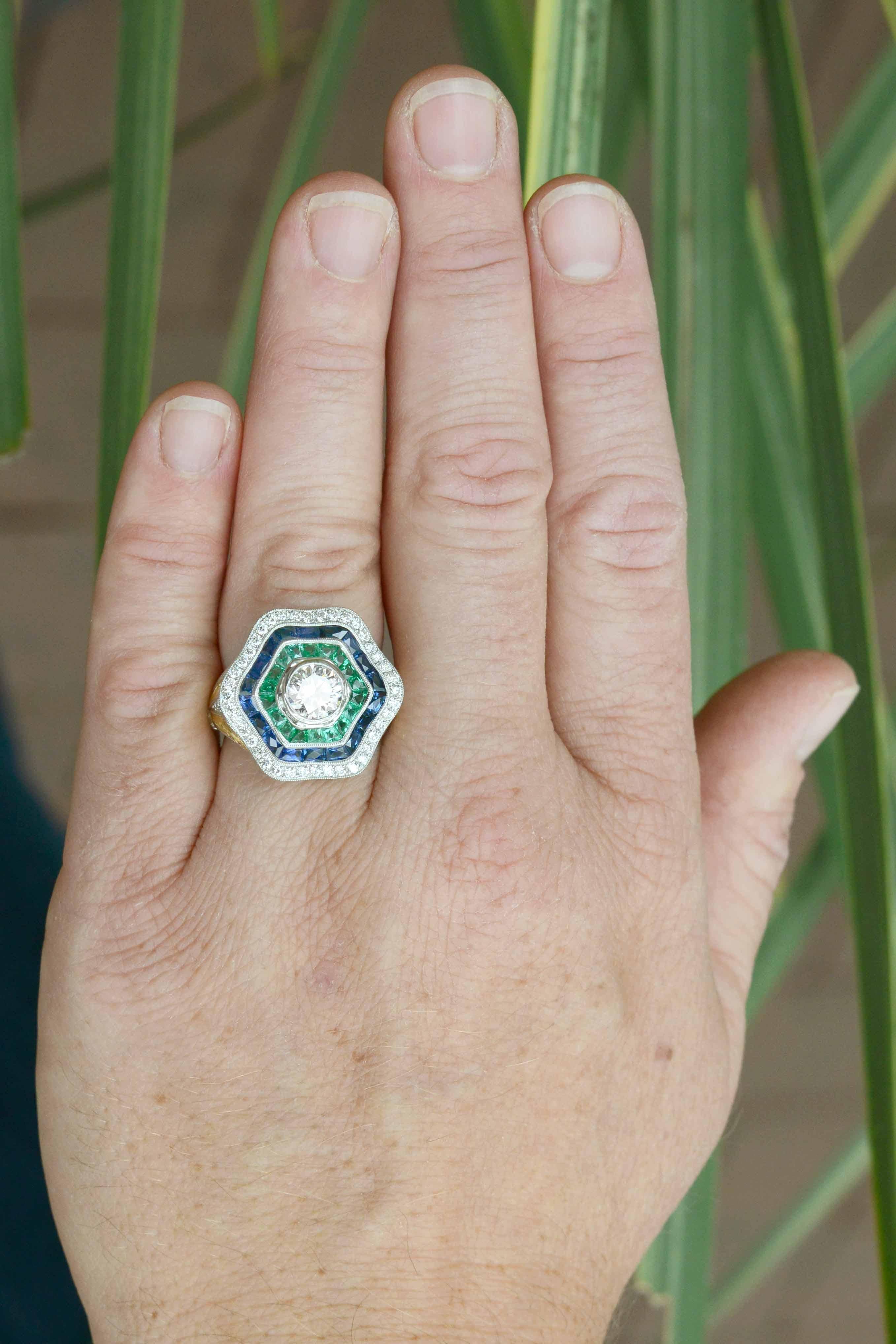 A dynamic Art Deco style diamond, sapphire and emerald ballerina ring. This dramatic cocktail ring is centered by a shimmering 1.02 carat round brilliant diamond bursting with fire. The undulating waves of calibre' and French cut gemstones resemble