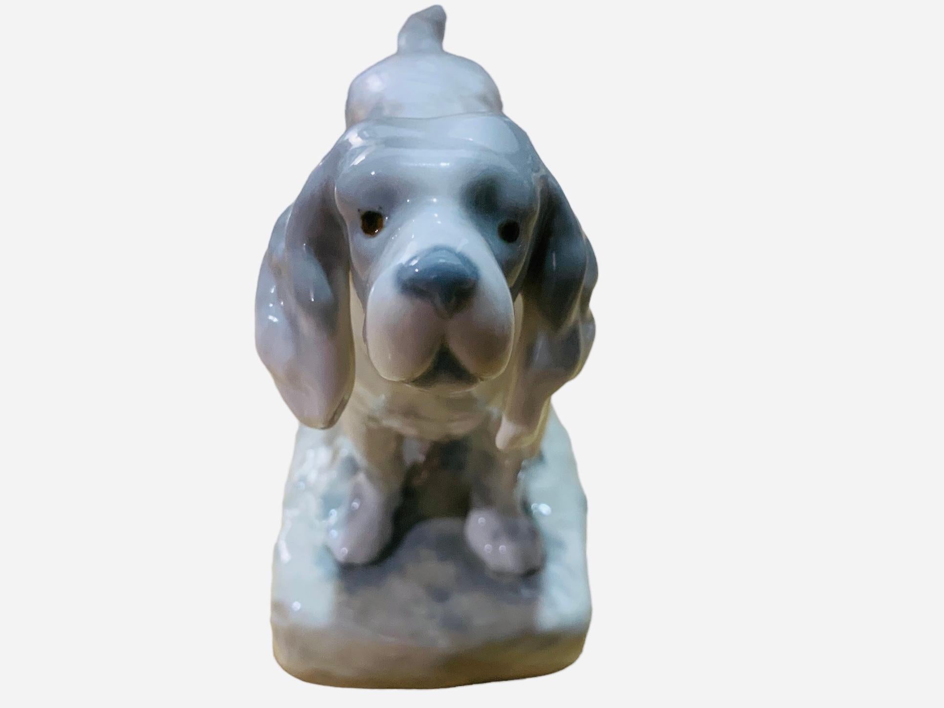 This is a Rex Valencia porcelain figurine of a dog. It depicts a hand painted grey and white color astute English Setter dog standing up over a base adorned with a cut trunk and soil. He has a beige belt in his neck, a straight tail and is ready to