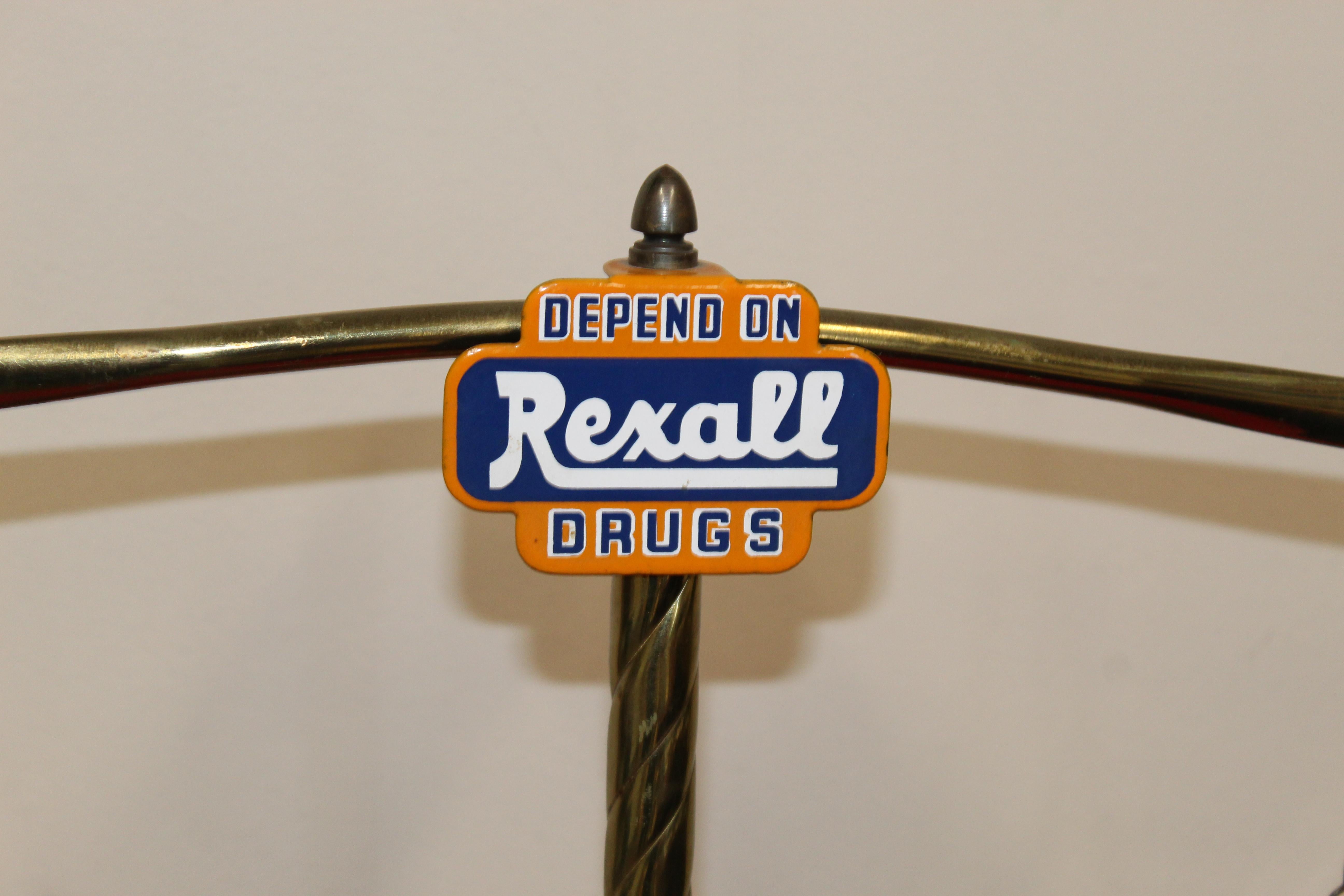 Counter top advertisement for Rexall Drugs, circa 1930s-1940s. It is a brass stand, designed to look like a set of balance beam scales with two round trays on chains attached to a rod that runs across the top. At the top decorated in blue and