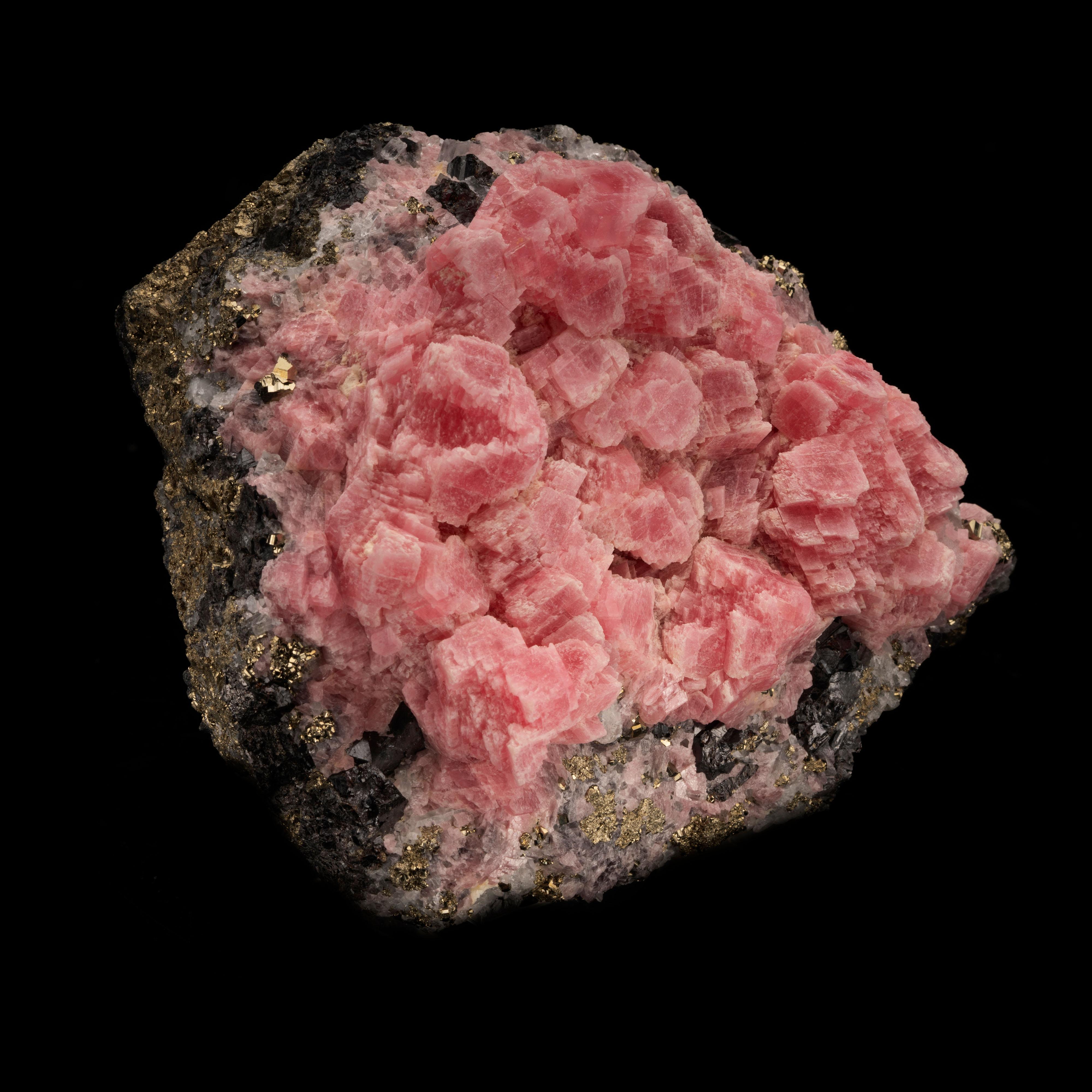 This stunning combination specimen out of Peru features lush pink rhombohedral rhodochrosite crystals densely clustered over a unique, sparkling matrix of lustrous, inky tetrahedrite and a generous coating of luxe pyrite crystals for a gilded finish