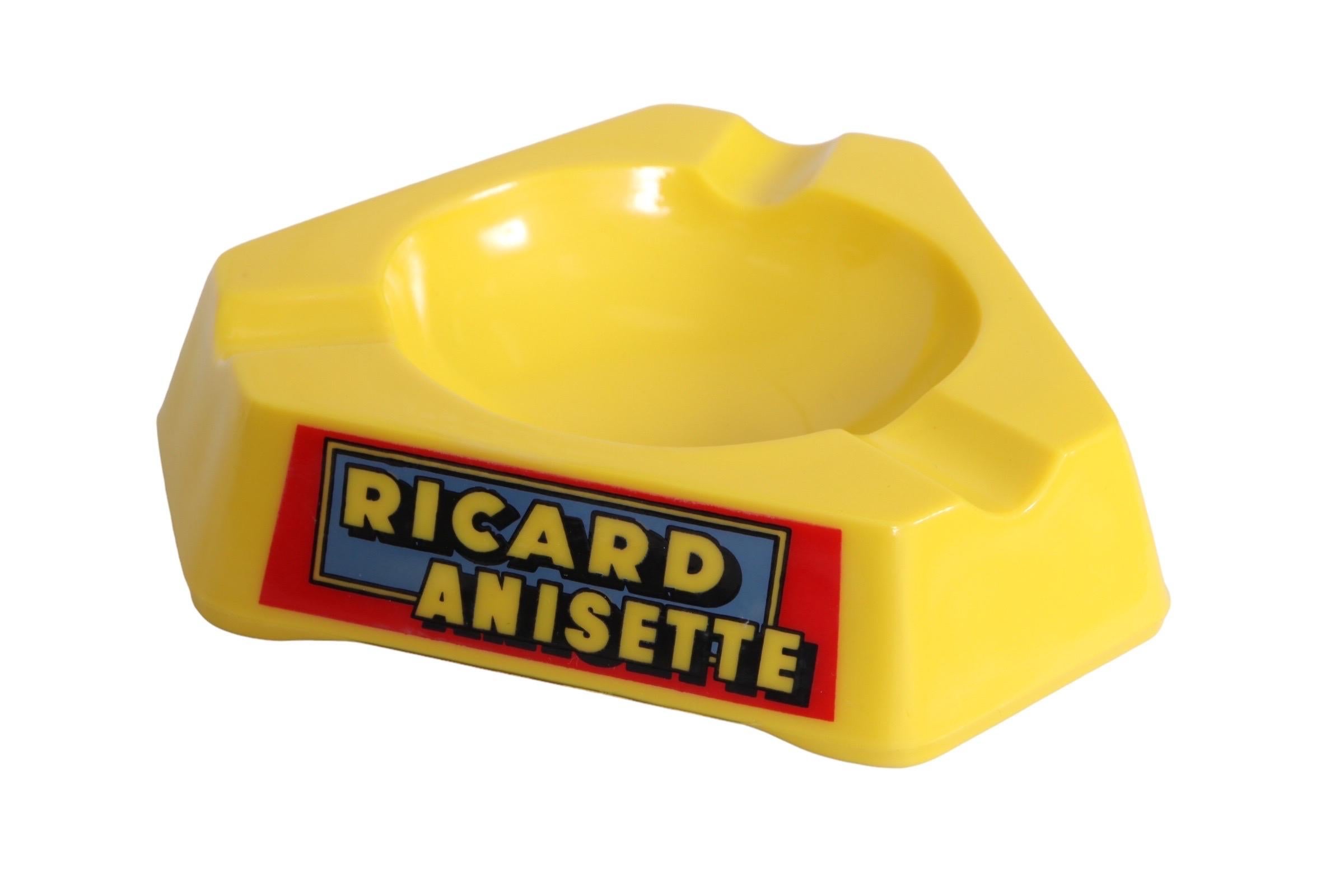 A Ricard branded triangular ashtray. Made of yellow opalex with a round central dish, each corner has beveled cigarette rests. Sides read Ricard Anisette in a bold font set over a blue and red background. Marked “Opalex, Made in France”