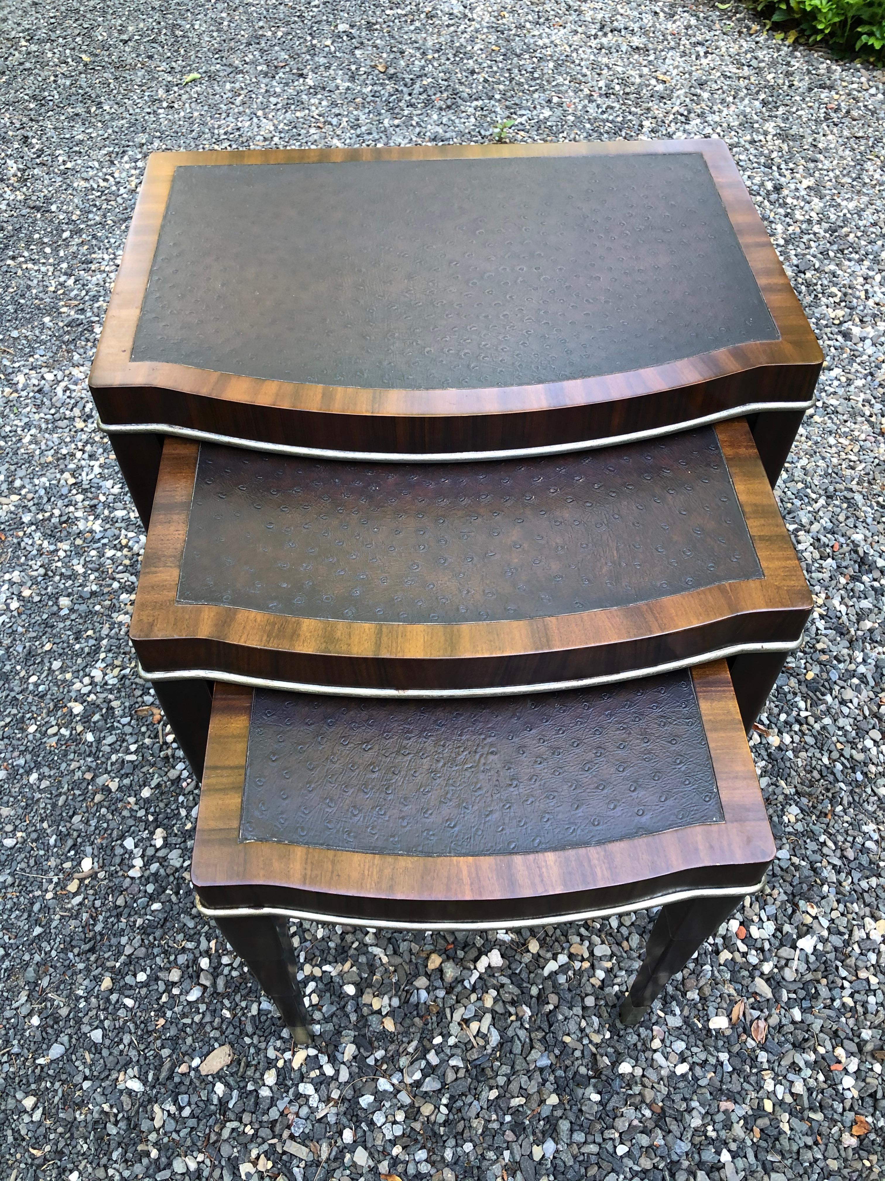 Set of three accent nesting tables in graduated sizes having mahogany structure and surrounds bordering a rich dark brown faux ostrich top. There is silver trim around the top and on feet. Only the largest table has cross bars at bottom. The legs on