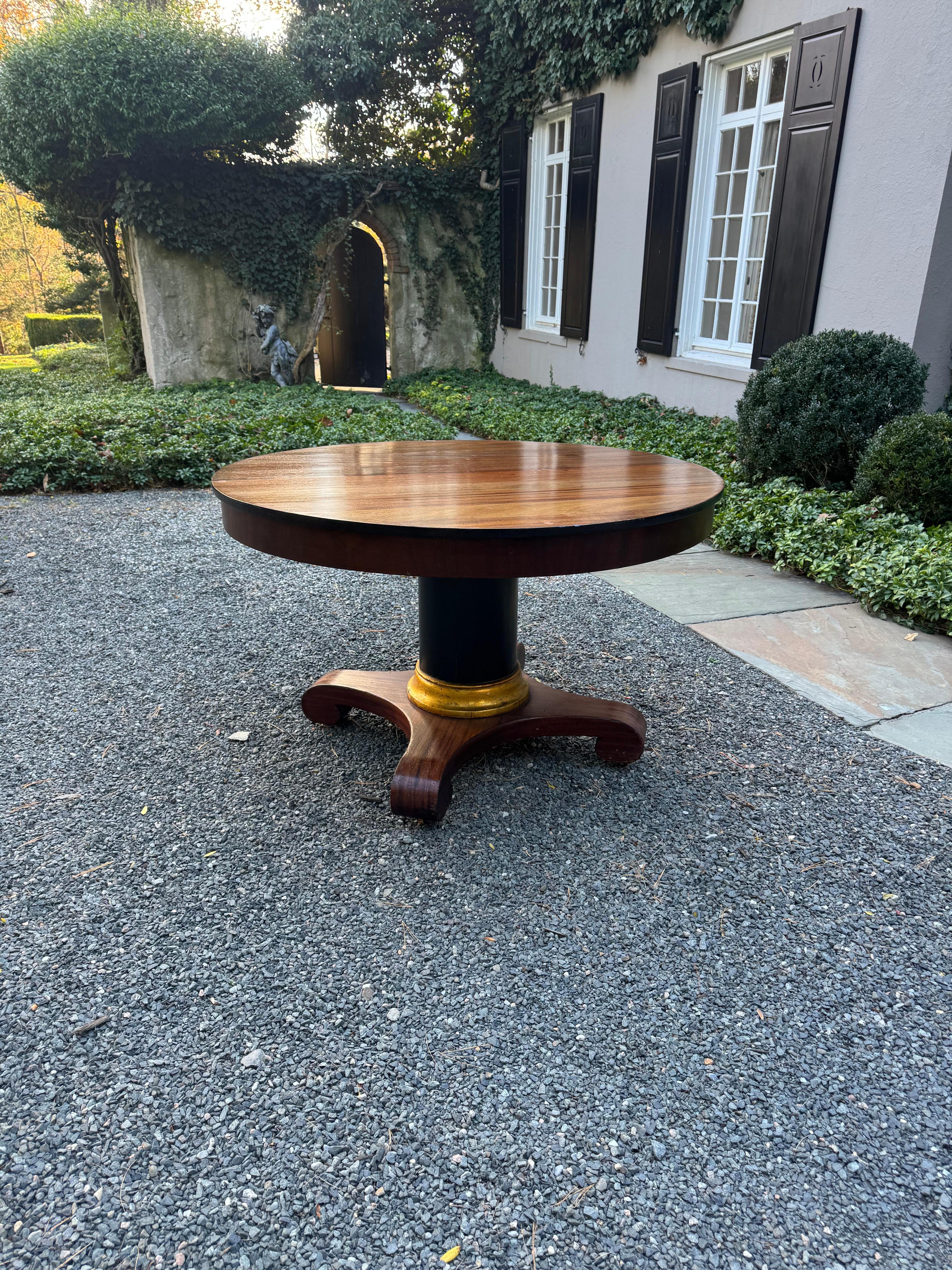 Table centrale ronde en acajou de l'époque Empire américain, avec un piédestal ébonisé et des détails dorés.  Structure solide avec des pieds tournés et des roulettes.  Un morceau de verre rond de 48 pouces est inclus si vous le souhaitez pour la