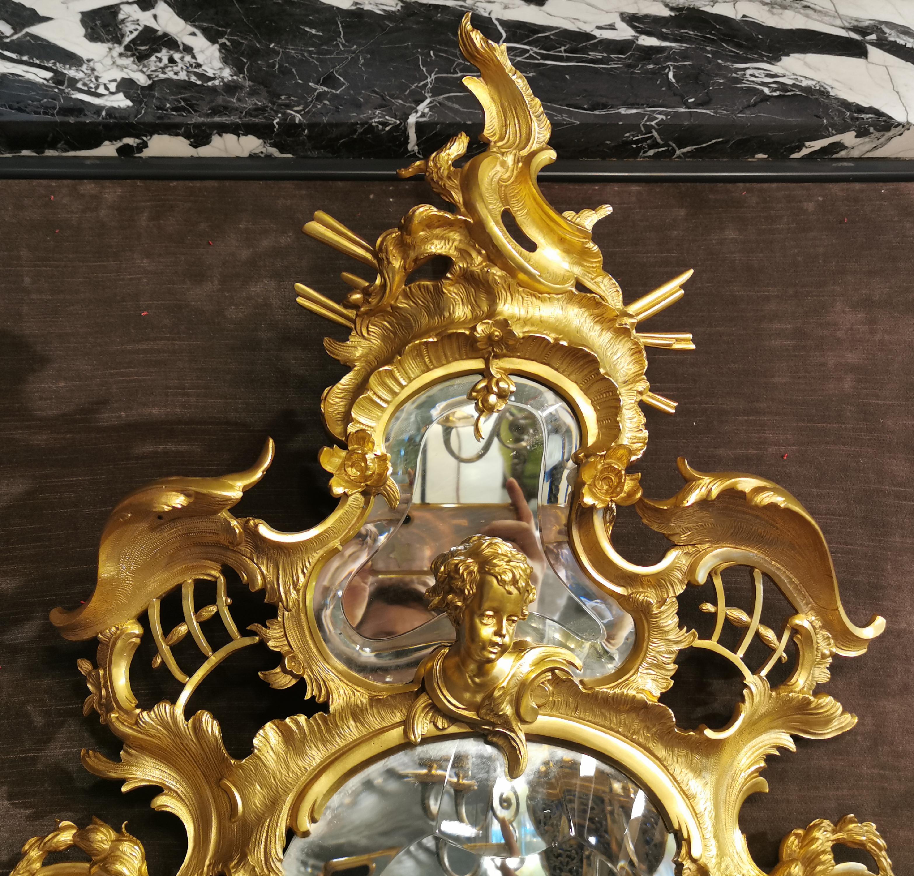 This very rich Napoleon III style mirror was made of gilt bronze in the second half of the 19th century.
The curved and countercurved frame with a very Rococo decoration frames two mirrors with beveled glass. In this decoration composed mainly with