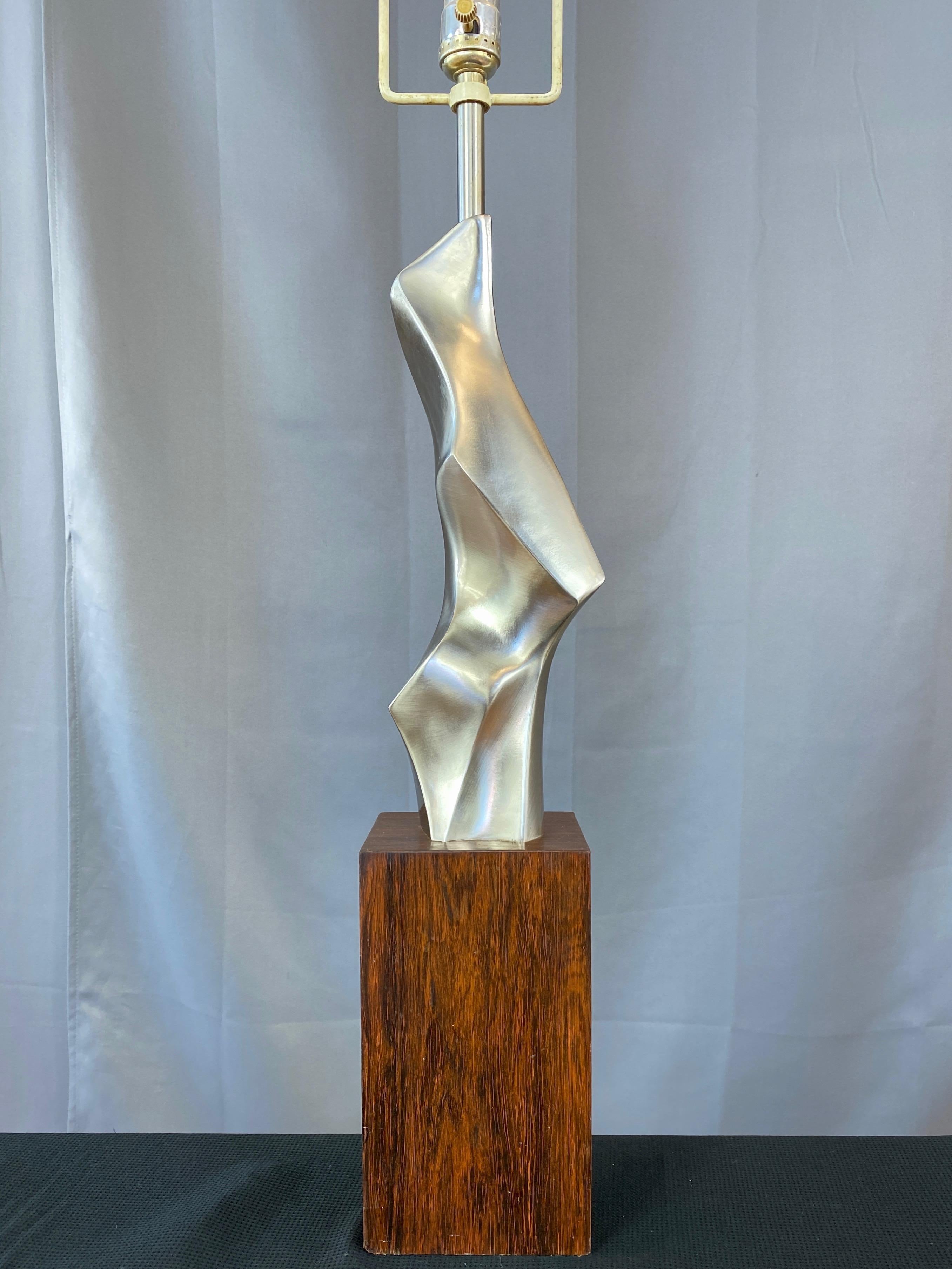 A fantastic 1963 mid-century modern abstract cast metal and rosewood table lamp by American designer Richard Barr for Laurel Lamp Company.

Artfully executed sculpture-like body of satin polished cast metal (appears to be aluminum or nickel) with