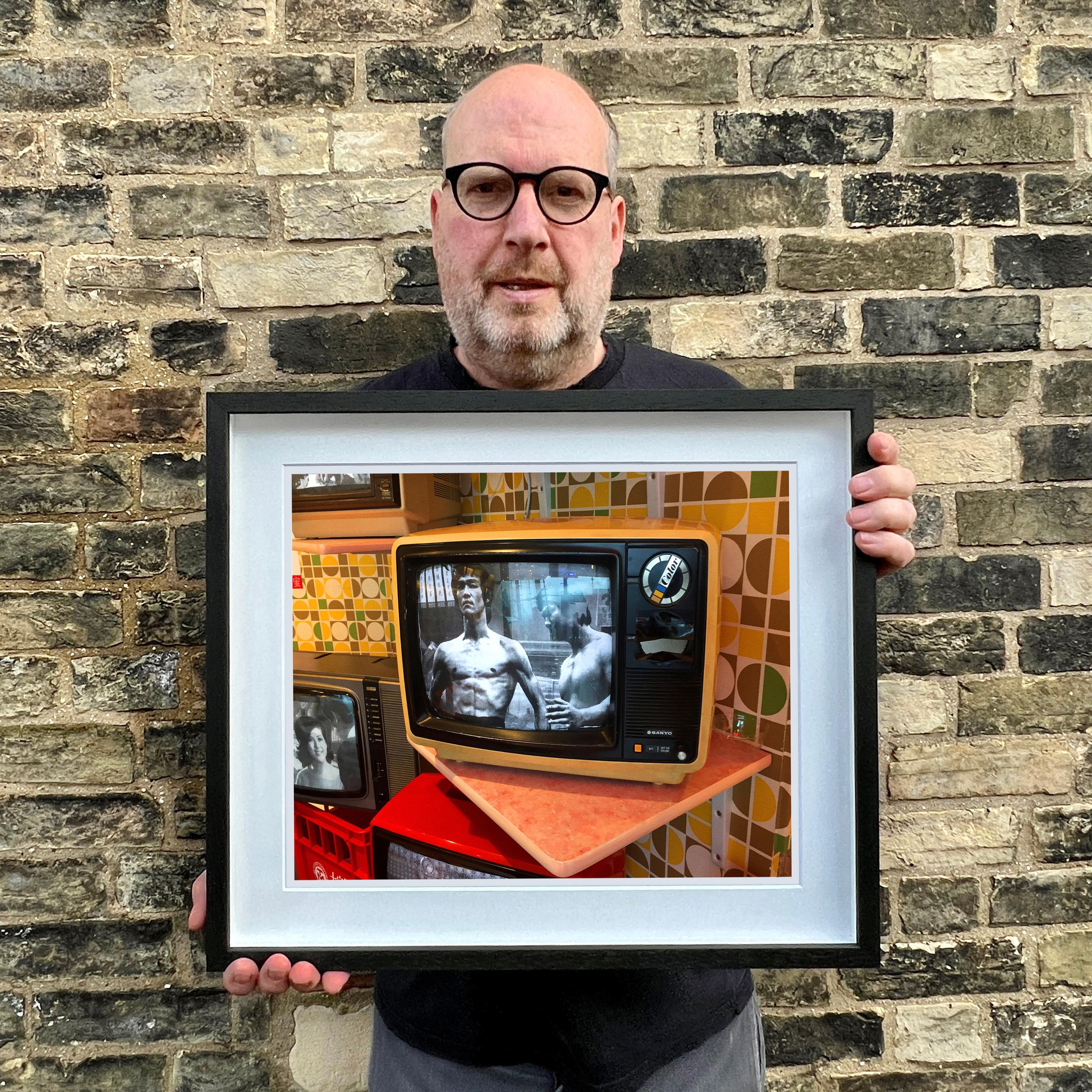 Bruce Lee TV, Foto von Richard Heeps, aufgenommen auf dem Gipfel des Victoria Peak in Hongkong. Eine von den siebziger Jahren inspirierte Senftapete bildet den Hintergrund für einen schwarz-weißen Vintage-Fernseher von Sanyo mit der Kampfsportikone