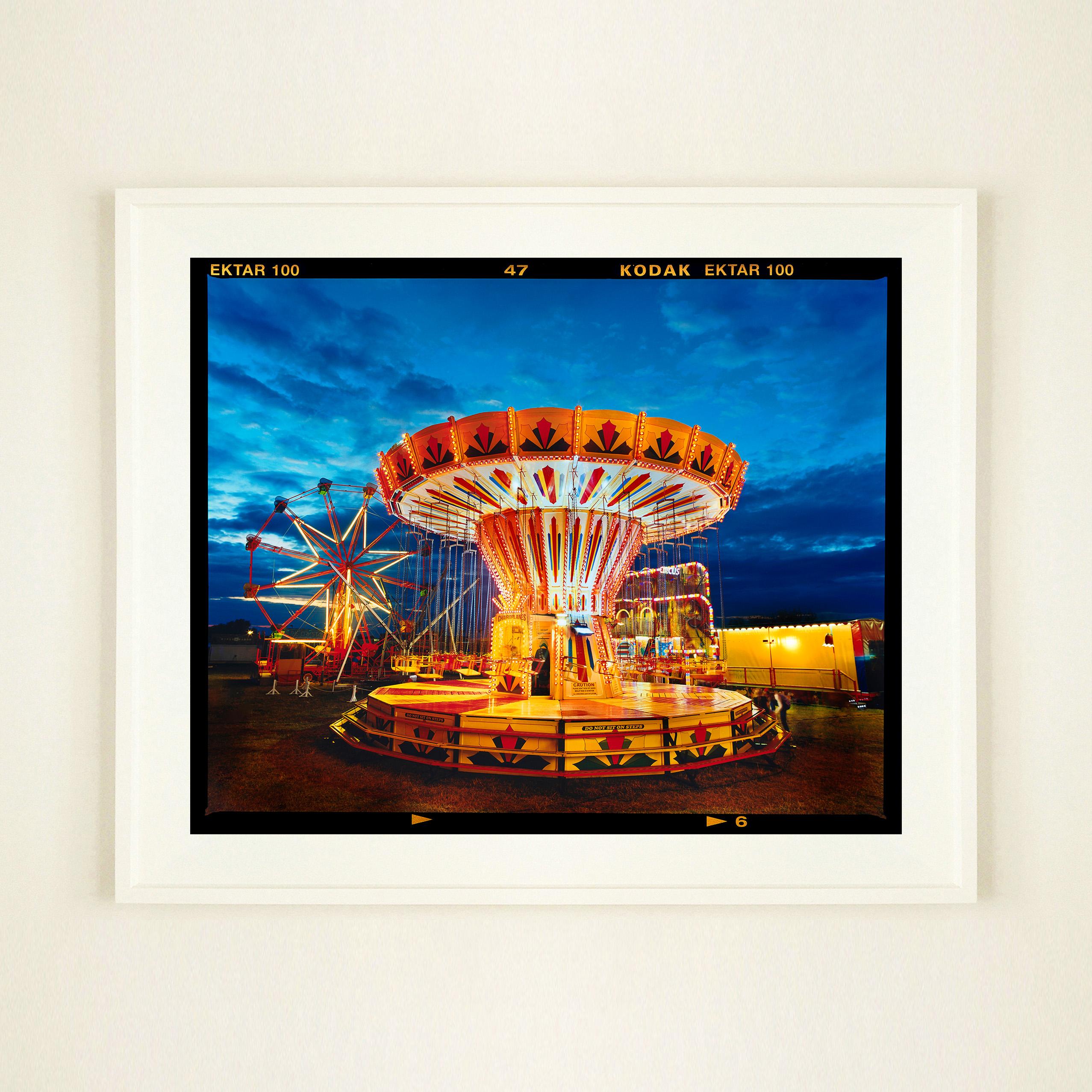 Photograph of a vintage fairground Chairoplane, lit up in a golden hue against the motion and beauty of the dark blue sky captures the magic of the 2022 Haddenham Steam Rally. Artwork from Richard Heeps autobiographical series, A View of the Fens
