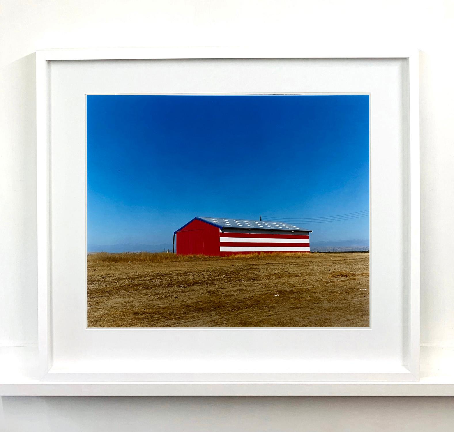 Stars and Stripes Barn, photography part of Richard Heeps Dream in Colour series. The American flag painted on a barn on the horizon, set against a big blue sky. 

This artwork is a limited edition of 25 gloss photographic print, dry-mounted to