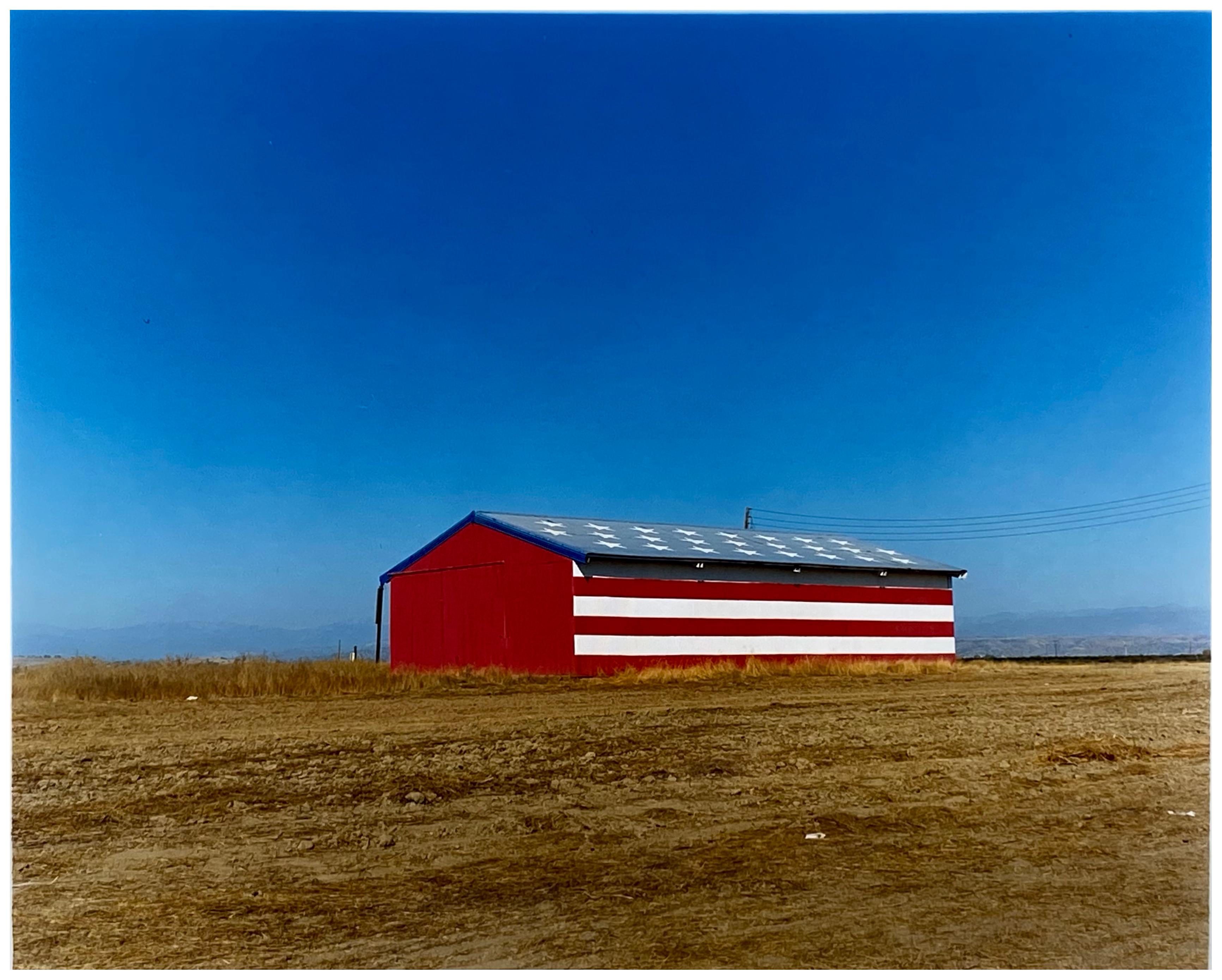 Stars and Stripes Barn, photography part of Richard Heeps Dream in Colour series. The American flag painted on a barn on the horizon, set against a big blue sky. 

This artwork is a limited edition of 10 gloss photographic print, dry-mounted to