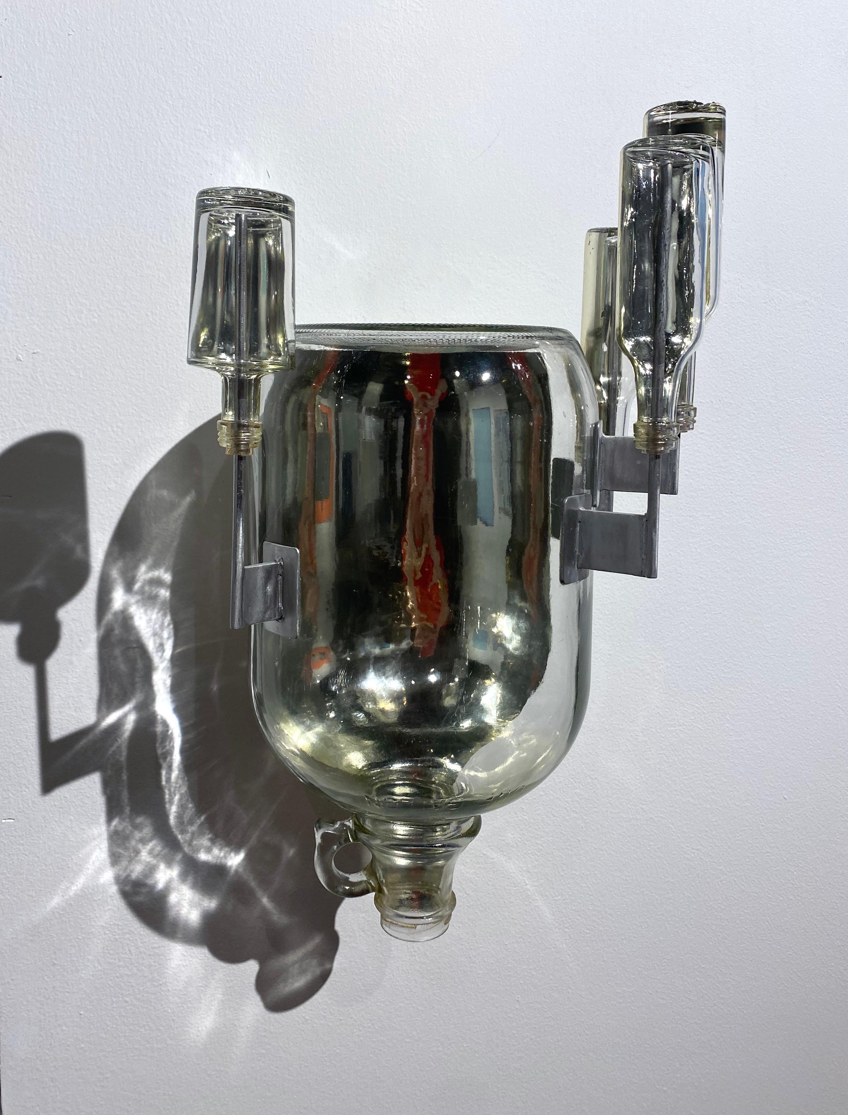 In this wall-mounted sculpture, glass bottles and a round, cylindrical jug are mirrored with white gold leaf on half the inside, creating a reflective inner side visible through the clear front.

Richard Klein has utilized found objects in his