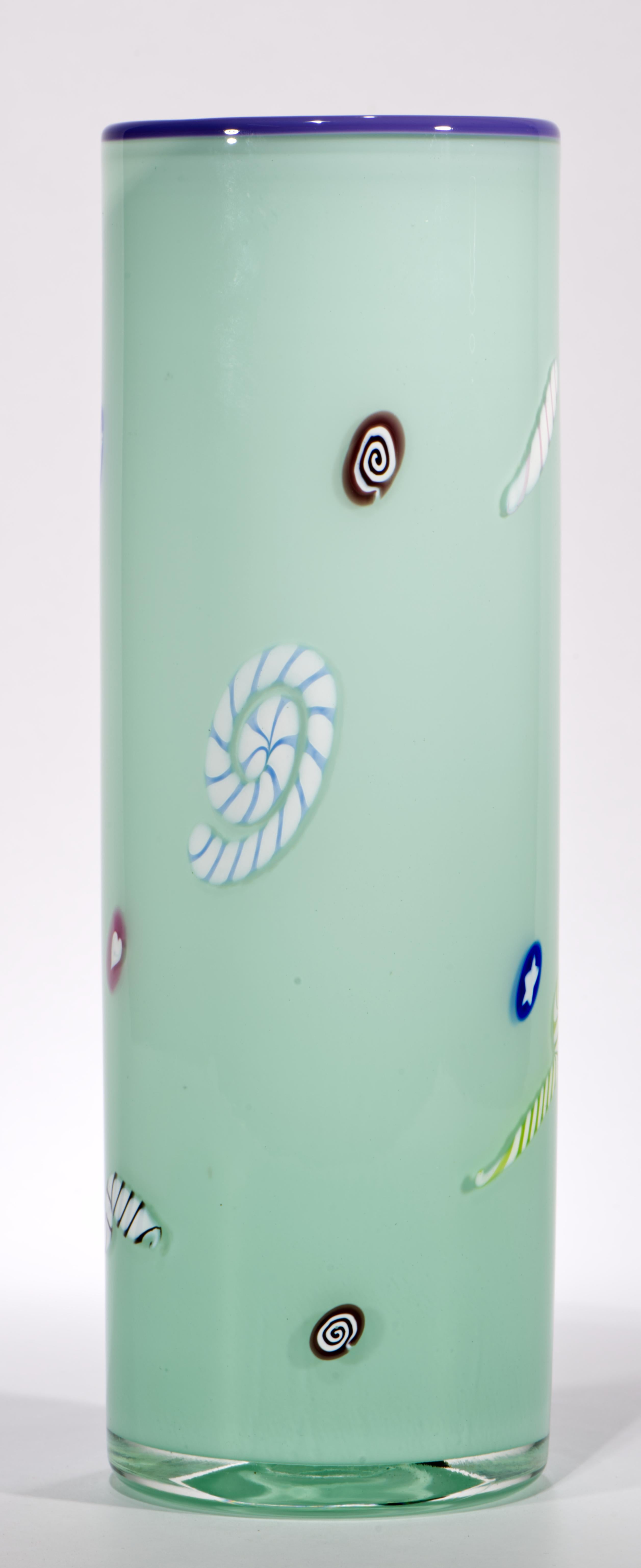 Il vaso in vetro artistico d'epoca ha un corpo cilindrico con decorazioni libere o grandi murrini e canne su uno sfondo blu celeste chiaro. Il bordo è accentuato da una fascia di vetro viola chiaro, mentre il corpo in vetro colorato è racchiuso in