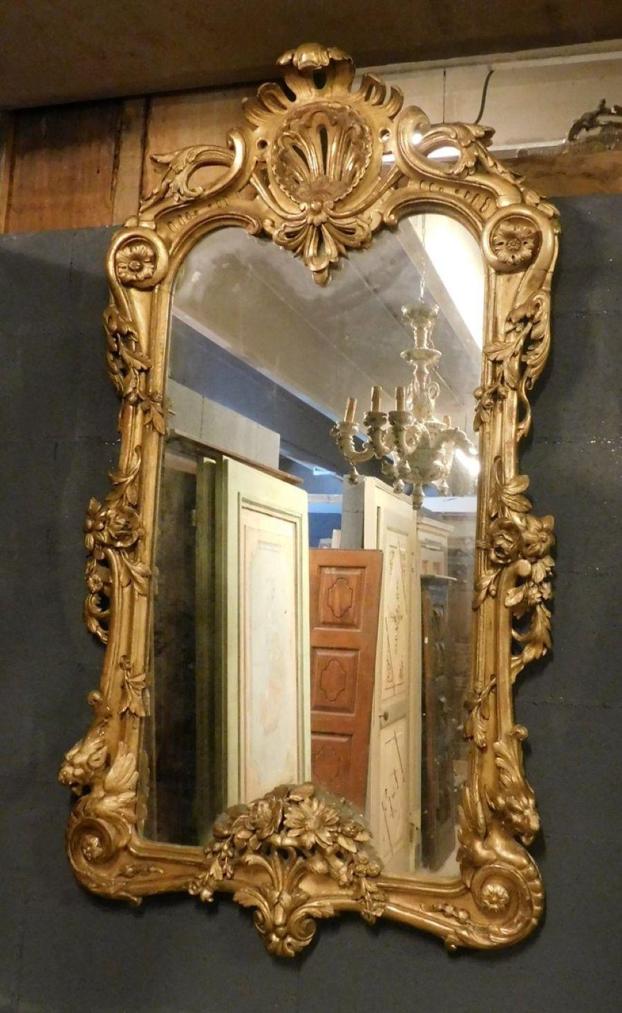 Antica Specchiera in legno intagliato e dorato, riccamente adornata con Decori a volute floreali e vegetali. Nella parte superiore è presente una ricca cimasa coronata centrata da conchiglia. La parte inferiore è arricchita con coppie di leoni alati