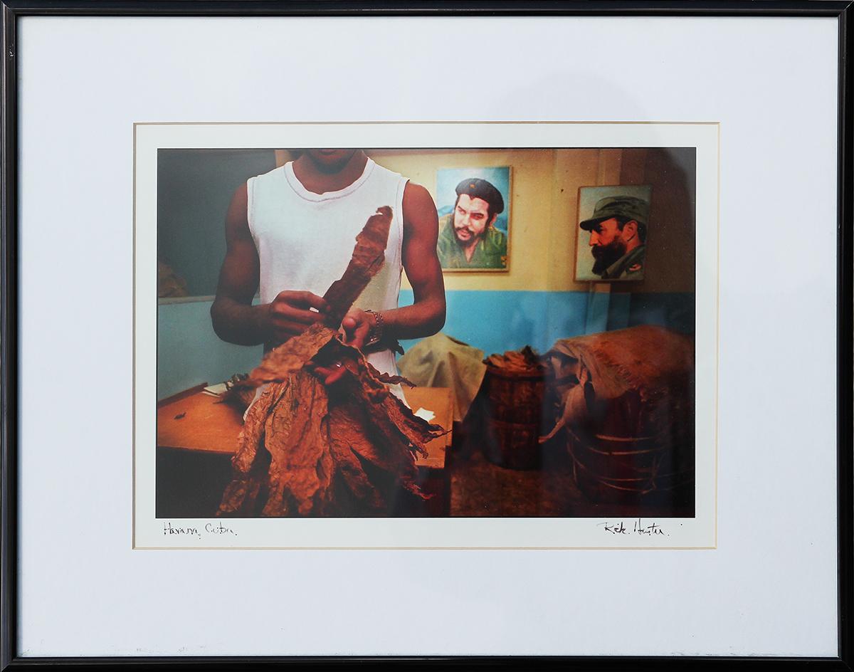 Photograph documenting daily life in Havana, Cuba by renowned photojournalist Rick Hunter. This work features a man working with tobacco leaves, likely to be rolled to produce cigars with a photo of Fidel Castro in the background. Signed by the