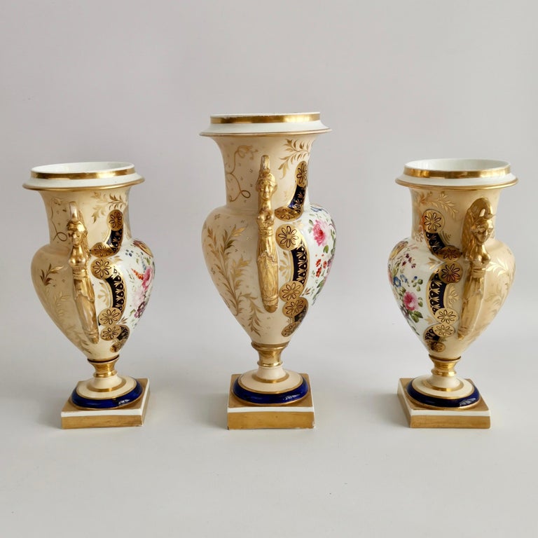 This is a spectacular garniture of three vases made by an English factory between 1810 and 1815. The vases are made in the French Empire style with heavily gilded Egyptian caryatid scroll handles, exceptionally beautiful flower reserves on the