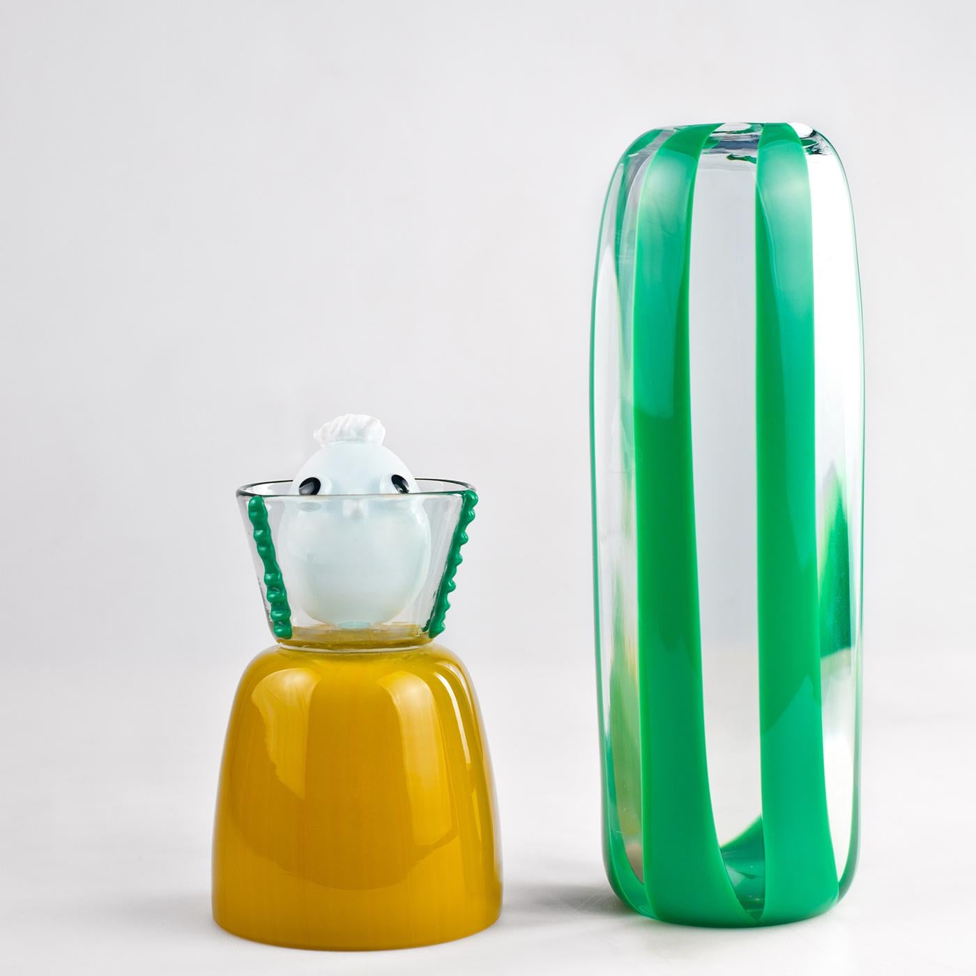 A unique character in three pieces for a mysterious effect. With a mustard yellow bust, the piece rests on a lively base in transparent glass with green stripes. The piece is topped with a transparent glass collar, protecting a white glass head with