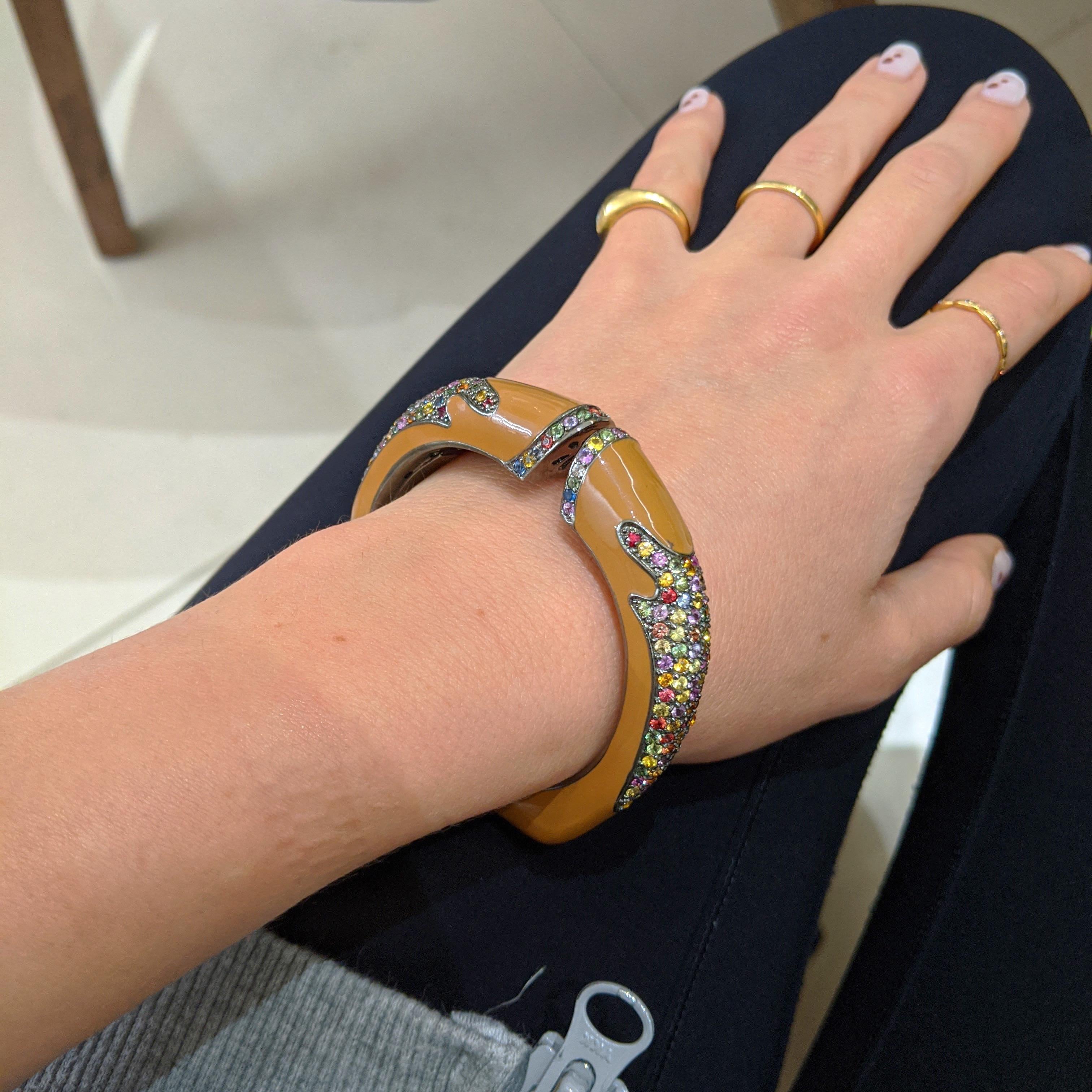 A caramel colored enamel bracelet set with multi colored round brilliant cut Sapphires. The back of the bracelet is Sterling Silver. The hinge is at the bottom of the bracelet.
The inside measurements are 2 1/4