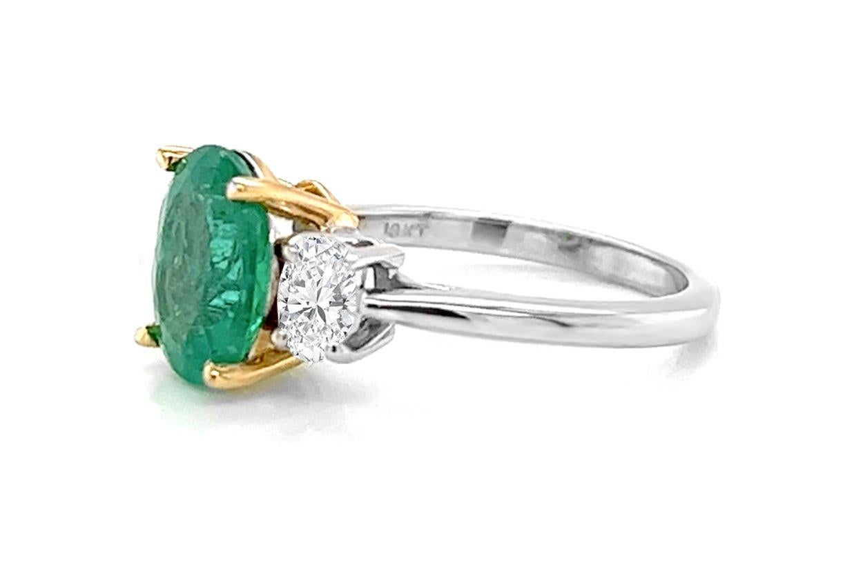 This 18kt gold ring showcases a stunning GIA-certified oval emerald at its center, exuding a rich green hue and natural beauty. Flanking the emerald on either side are sparling oval diamonds, which complements the emerald's brilliance and add a