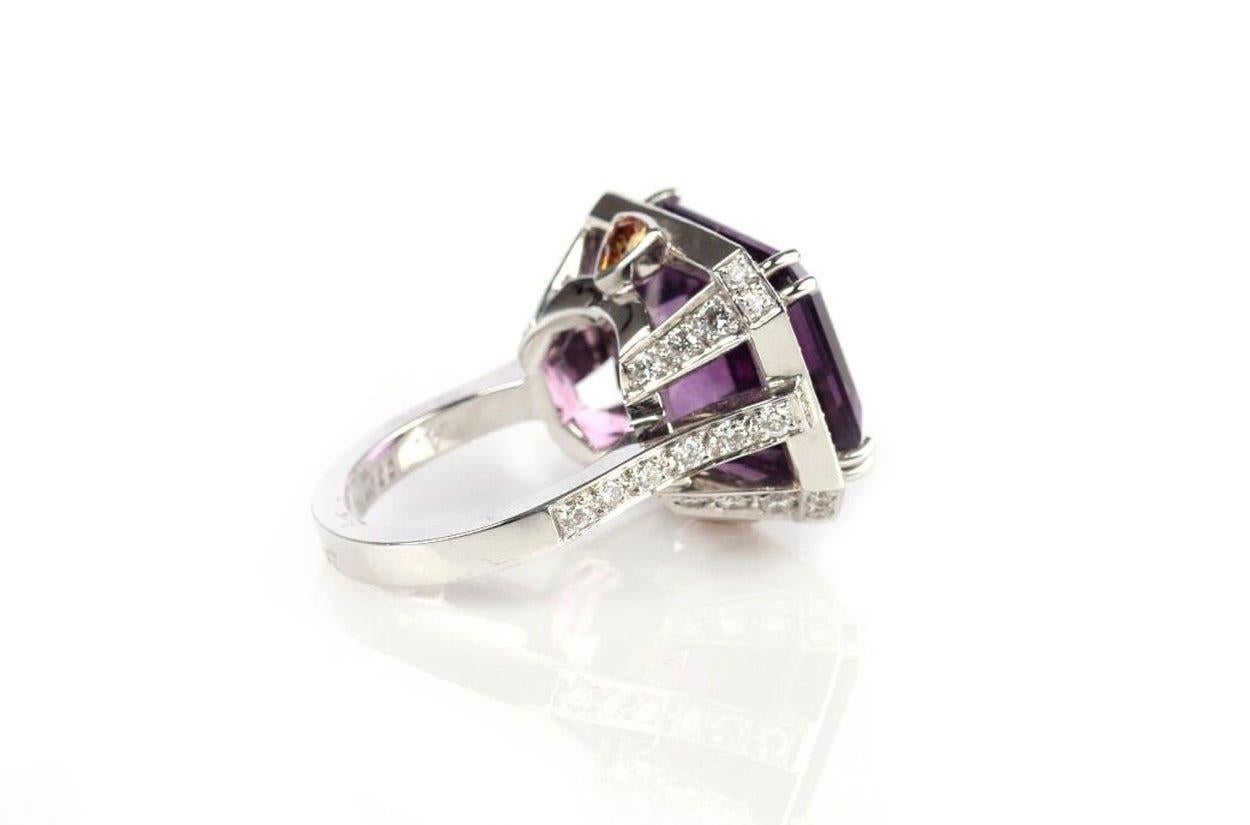 A splendid ring 18kt white gold showcases a mesmerizing center emerald-cut amethyst, which exudes an air of regal elegance. Encircling this captivating amethyst are vibrant orange round sapphires, creating a striking contrast that adds a touch of