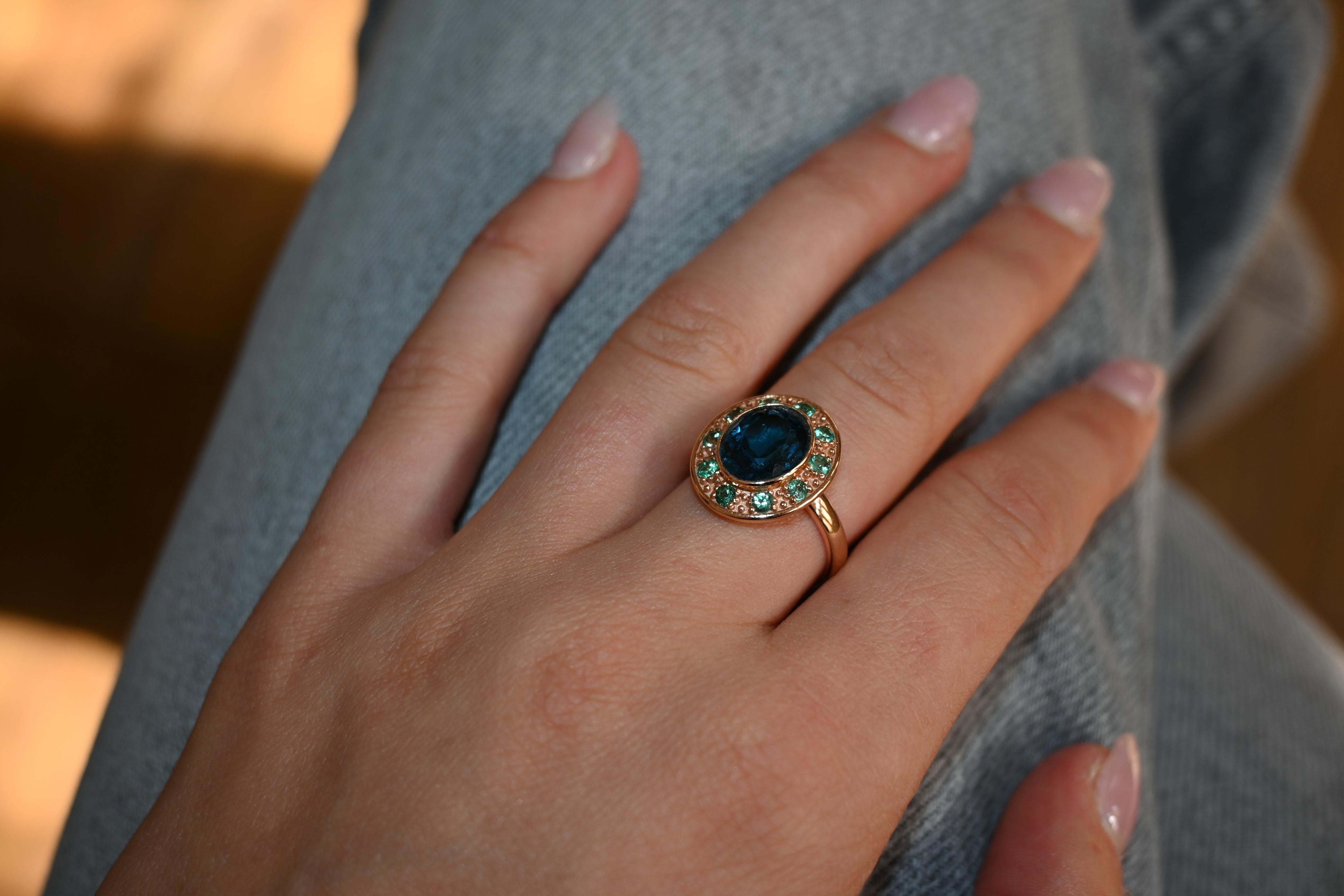 Anillo Topacio Azul Esmeralda Oro Rosa. 

En el centro del anillo hay un brillante topacio azul de 1,3 cm de diámetro. Esta piedra llama inmediatamente la atención, con diez pequeñas piedras verdes dispuestas a su alrededor y realzadas por finos