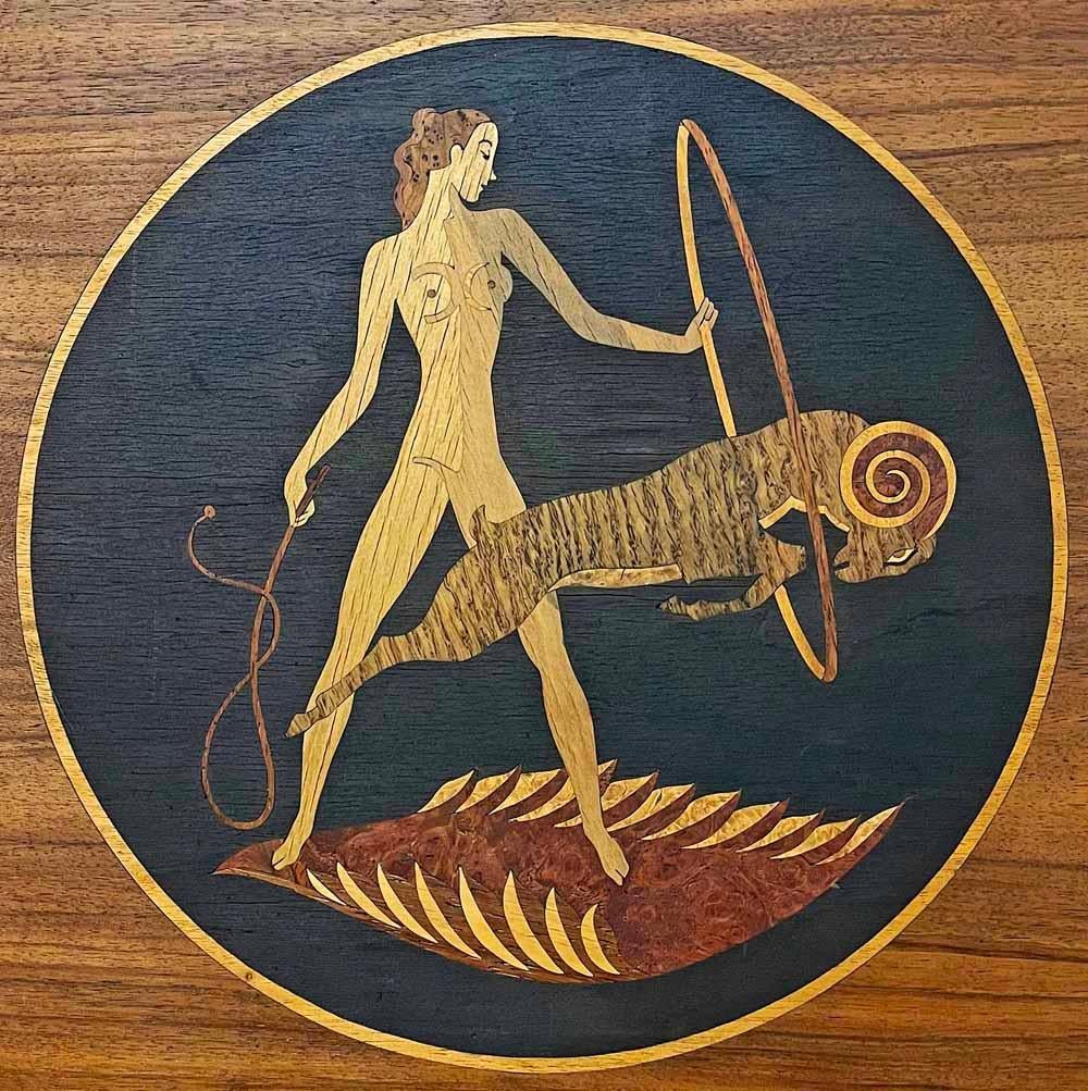 This masterwork of inlaid wood -- in stunning, interlocking pieces of burled walnut and maple, mahogany and beech -- depicts a nude female ring master with whip in hand, urging a ram to jump through her ring. The artist was Andrew Szoeke, who was