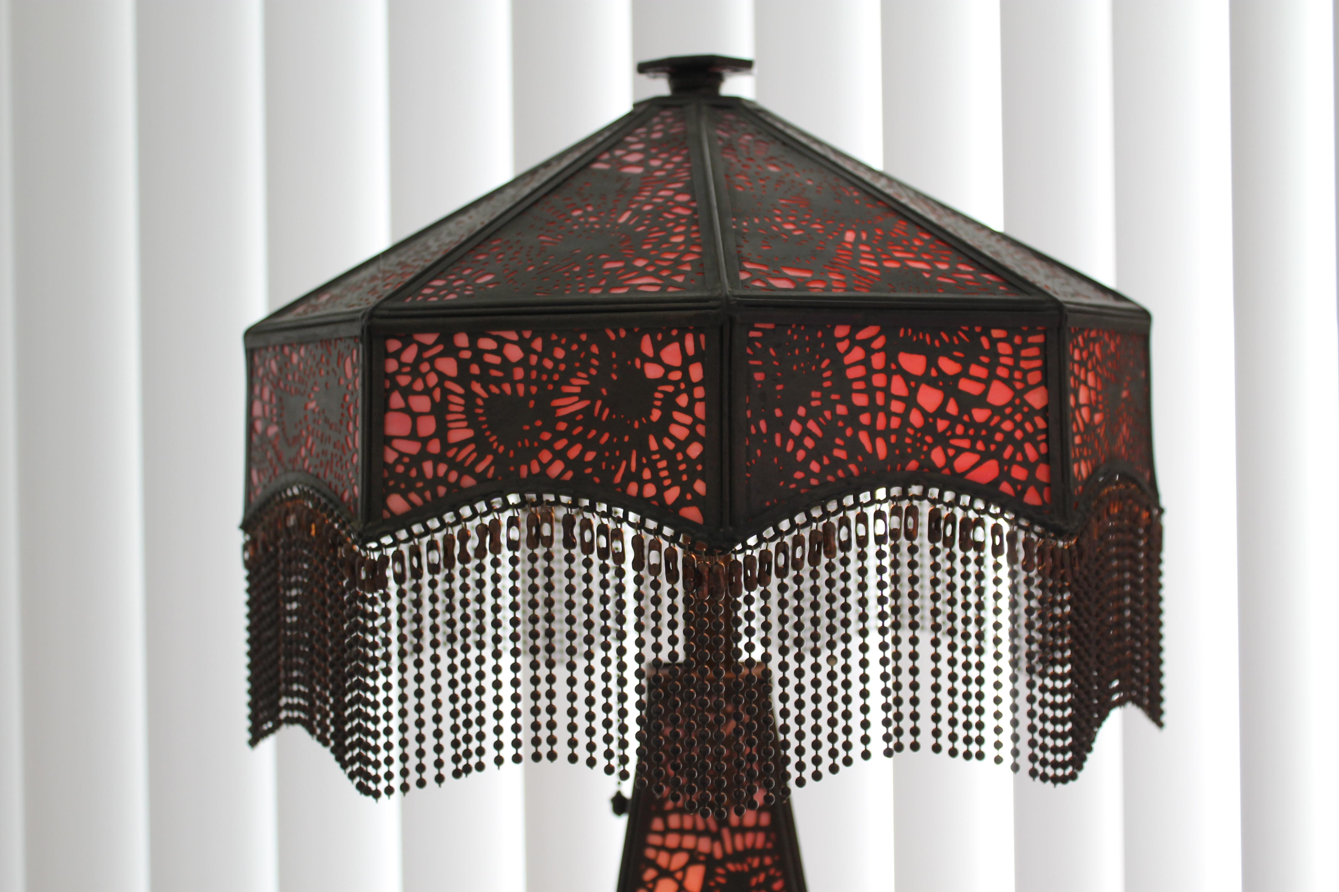 A circa 1920s table lamp with openwork bronze and rare rose colored glass. (Riviere Studios almost always used green glass) The base also lights and the lamp was recently rewired. Copper bead fringe is not original. All glass is intact with no