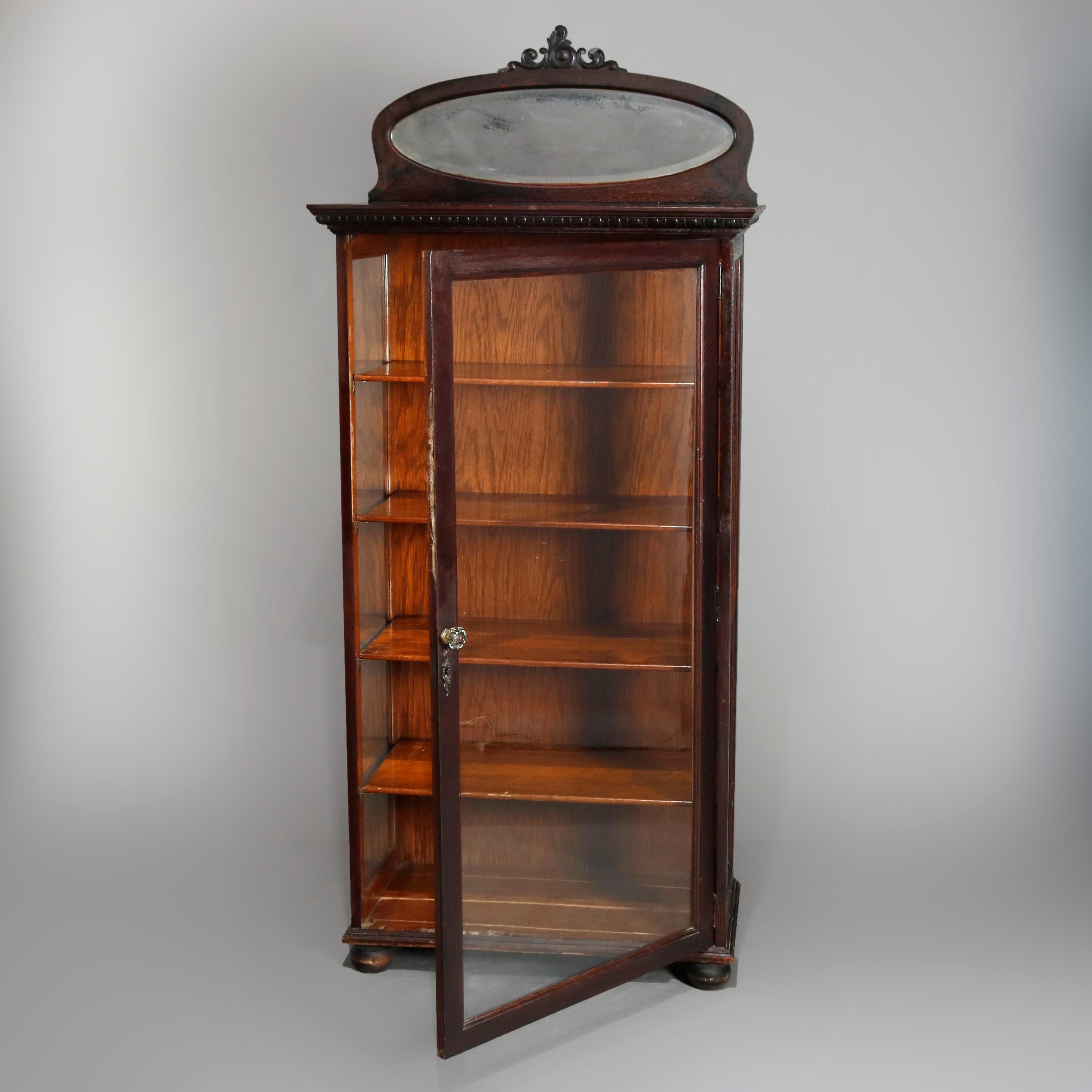 An antique R.J. Horner School china cabinet offers oak construction with backsplash with oval frame having carved foliate crest and beveled mirror surmounting case with single glass sides and door opening to shelved interior and raised on ball feet,