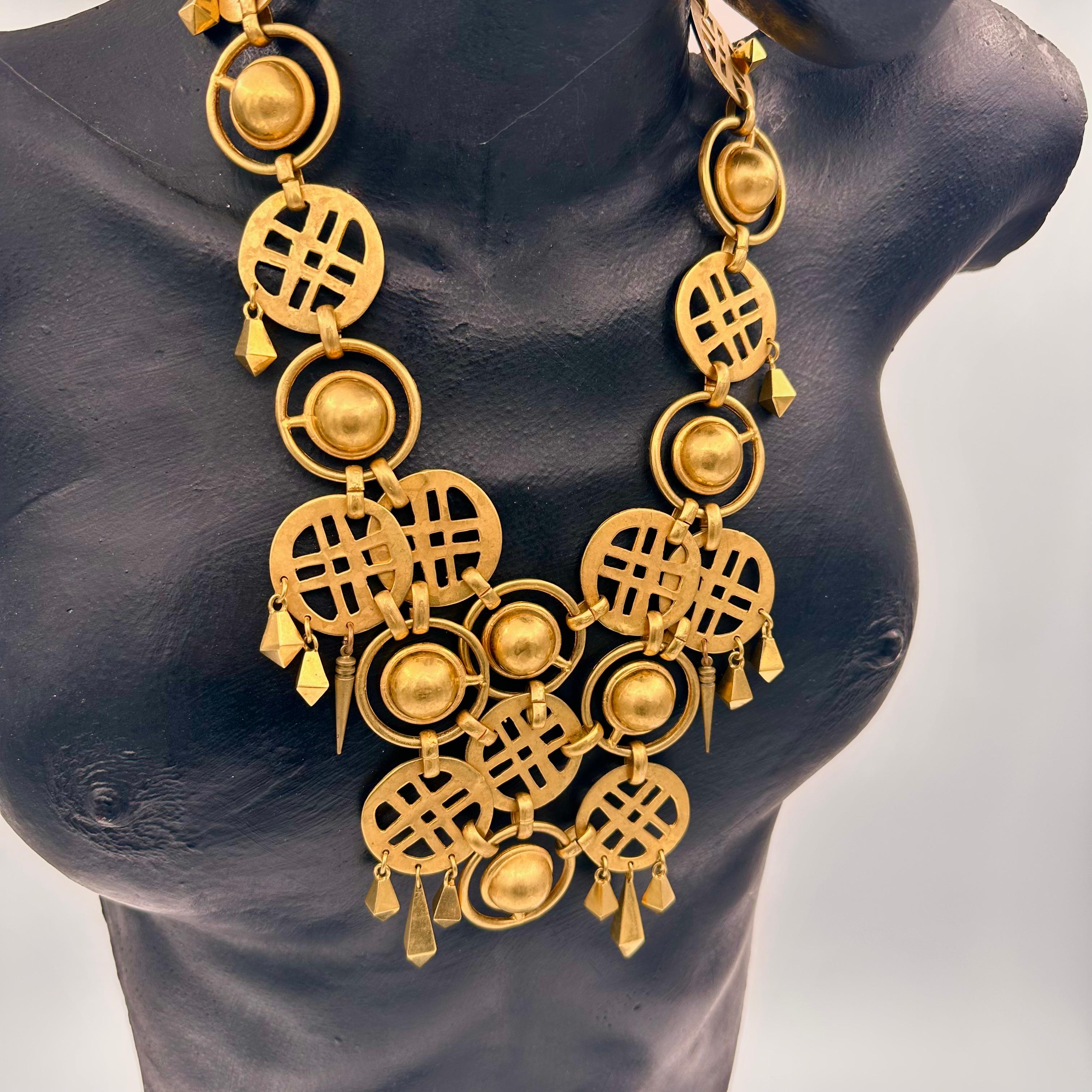 This is a configurations of gold plated brass crosshatched designs within circles and high domes surrounded by an orbiting ring, with chiseled fringe drops of solid brass, patterned into a high neck, short bib necklace. Simple hook clasp. One of a