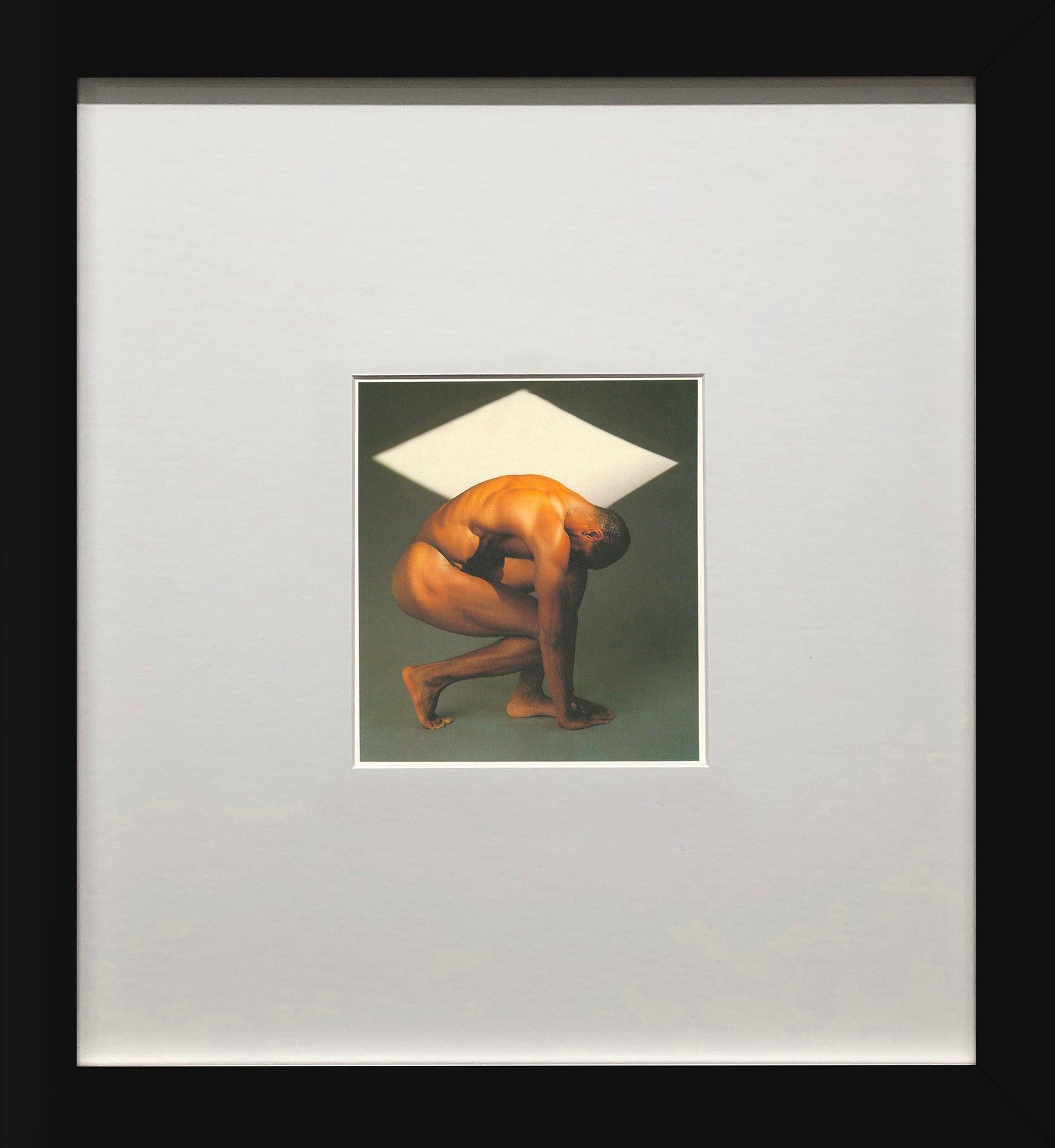 Cette image évocatrice de Robert Mapplethorpe met en scène le modèle Thomas, capturé dans une pose gracieuse, légèrement accroupi. Sur un fond neutre ponctué d'une forme géométrique blanche comme un diamant, la composition crée un contraste visuel