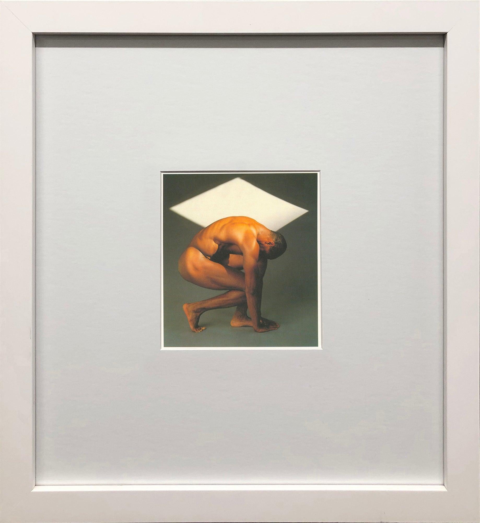Cette image évocatrice de Robert Mapplethorpe met en scène le modèle Thomas, capturé dans une pose gracieuse, légèrement accroupie. Avec une toile de fond neutre accentuée par une forme géométrique blanche comme un diamant, Mapplethorpe crée un