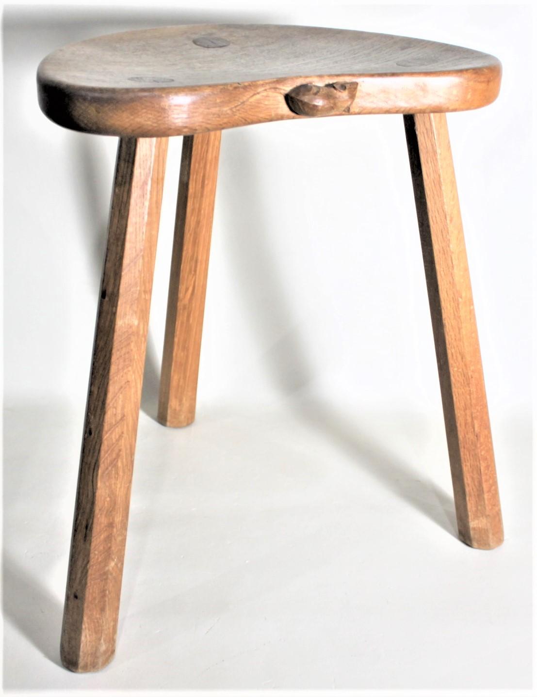 This vintage solid oak three legged milking stool was made by the renowned English furniture maker Robert 'Mouseman' Thompson in his signature folk art style and dates from circa 1960. The stool has a kidney shaped solid oak top with a slight