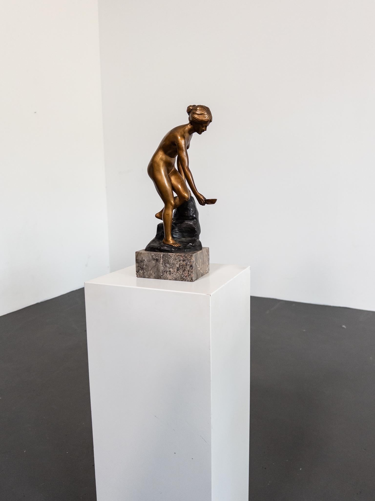 A finely detailed bronze sculpture by Austrian artist Robert Rudolfi (1873–1935), titled An der Quelle (“At the Spring”). The piece depicts a young woman gracefully gathering water in a shallow bowl, poised in a moment of quiet beauty and classical