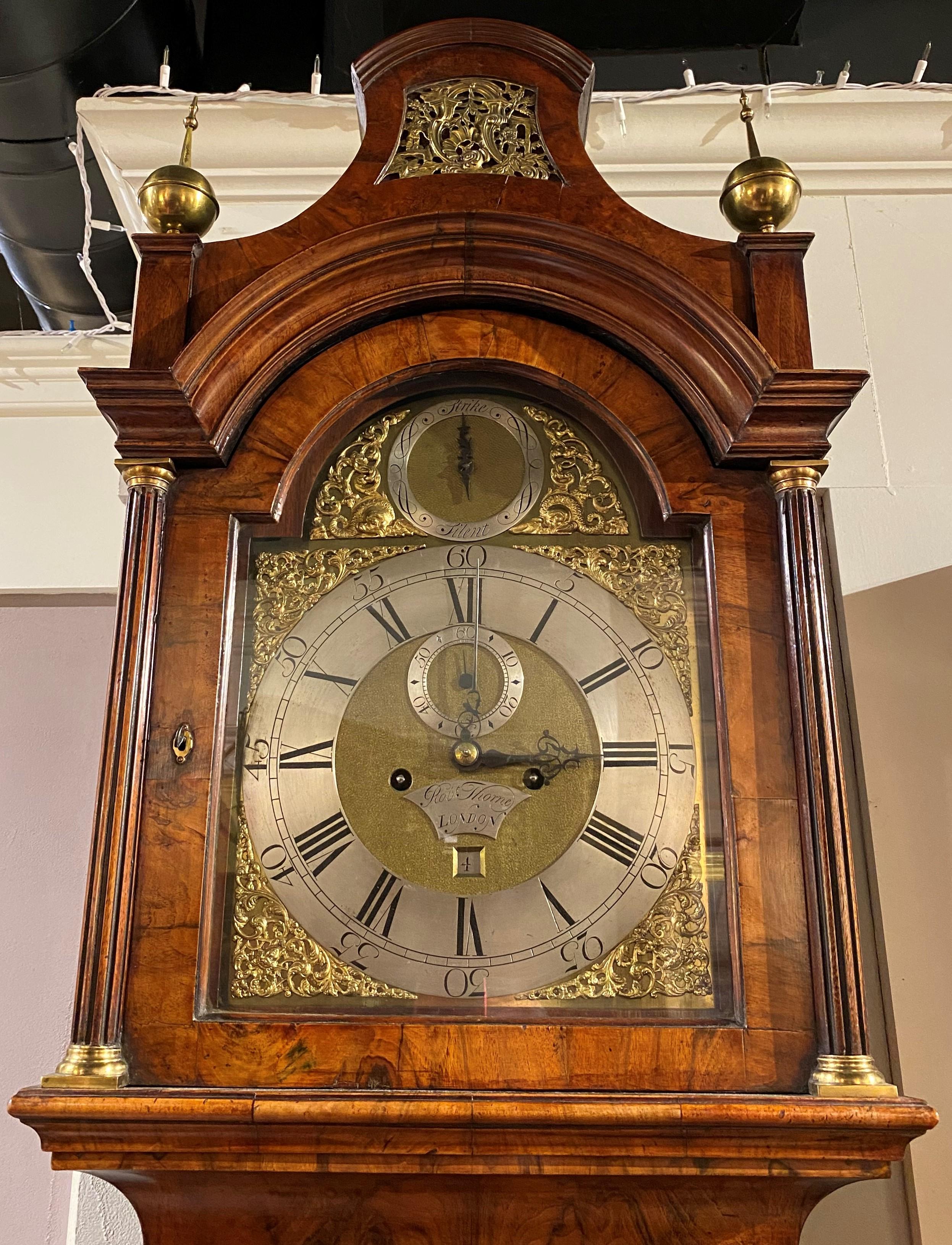 A stunning burled walnut tall case clock with pagoda form bonnet featuring a pierce-carved gilt central panel flanked by two ball and spire brass finials, surmounting an arched glass door, opening to an arched chased metal clock face with Roman